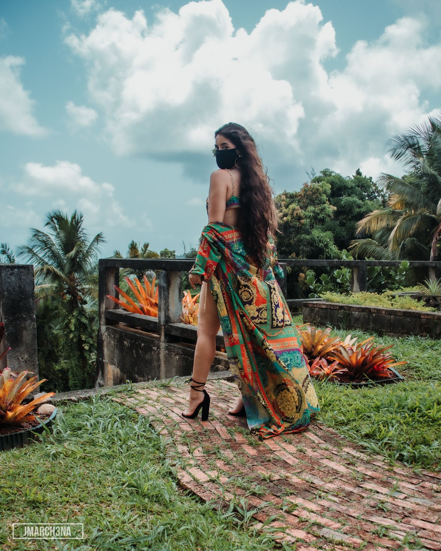 Woman in colorful maxi skirt and black mask poses on brick path surrounded by tropical greenery.