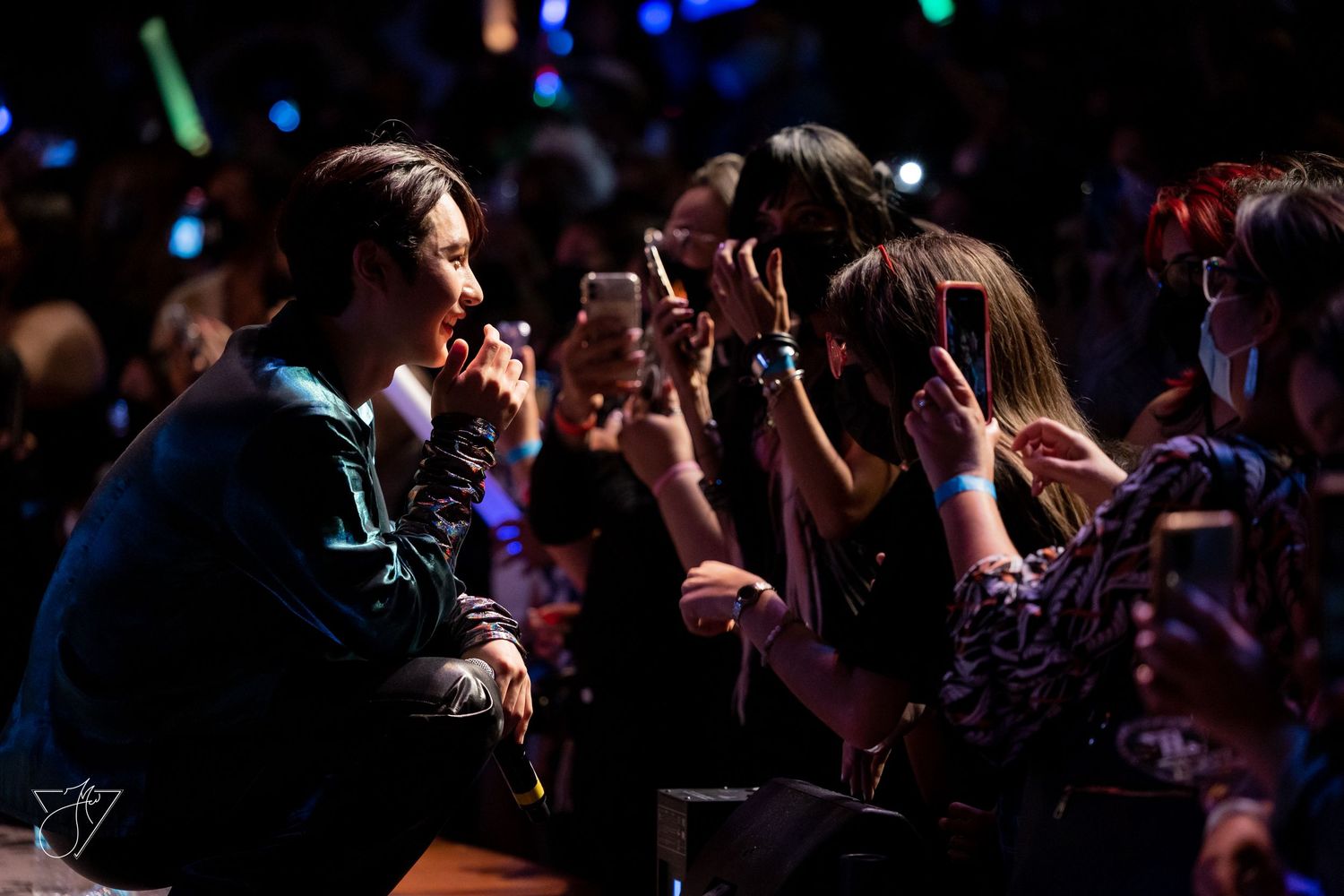 A performer interacts with fans holding phones at a concert, with colorful lights in the background.