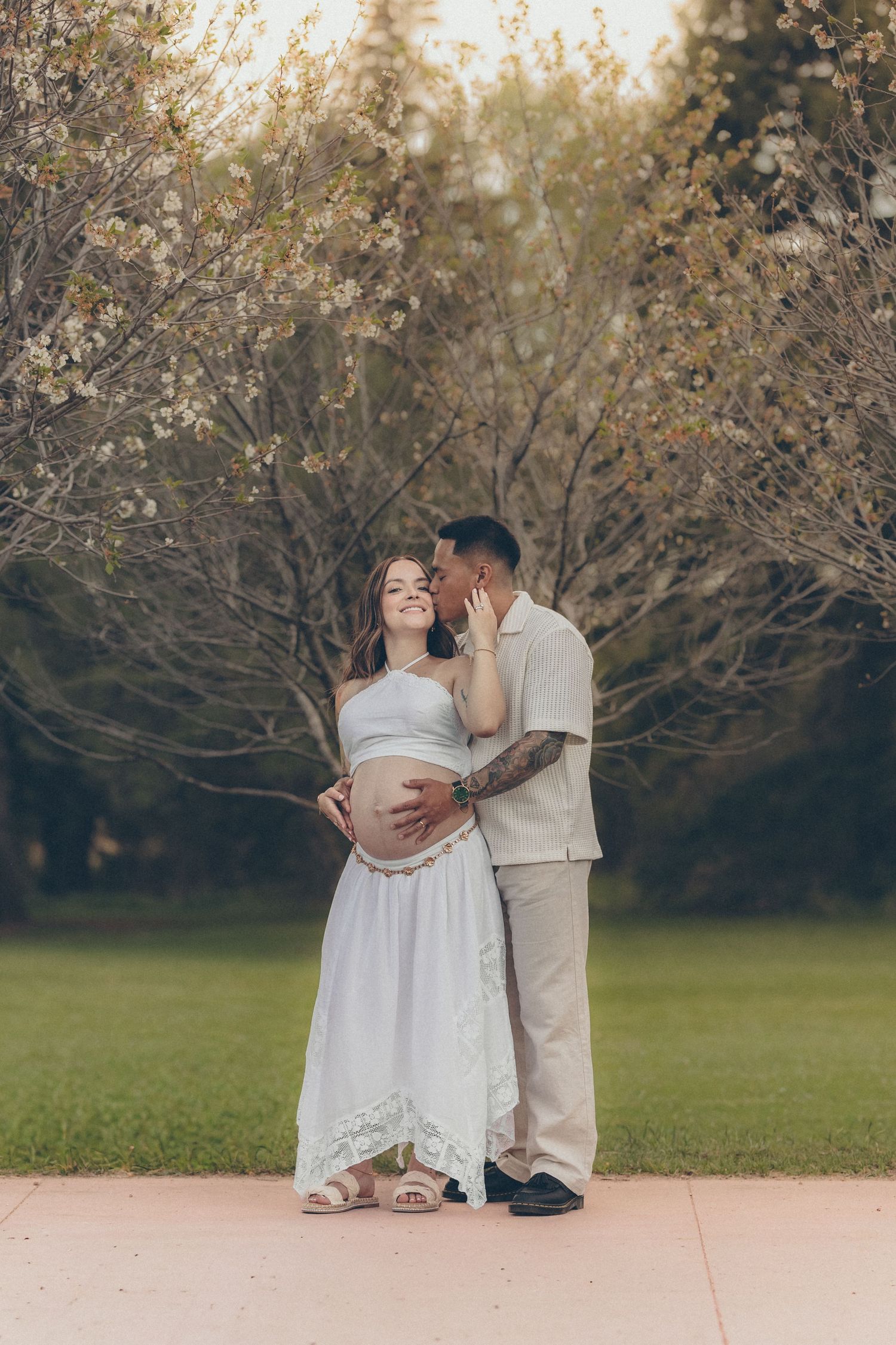 Couple sharing a tender moment outdoors, pregnant woman in white dress, man in beige outfit, blooming trees background.