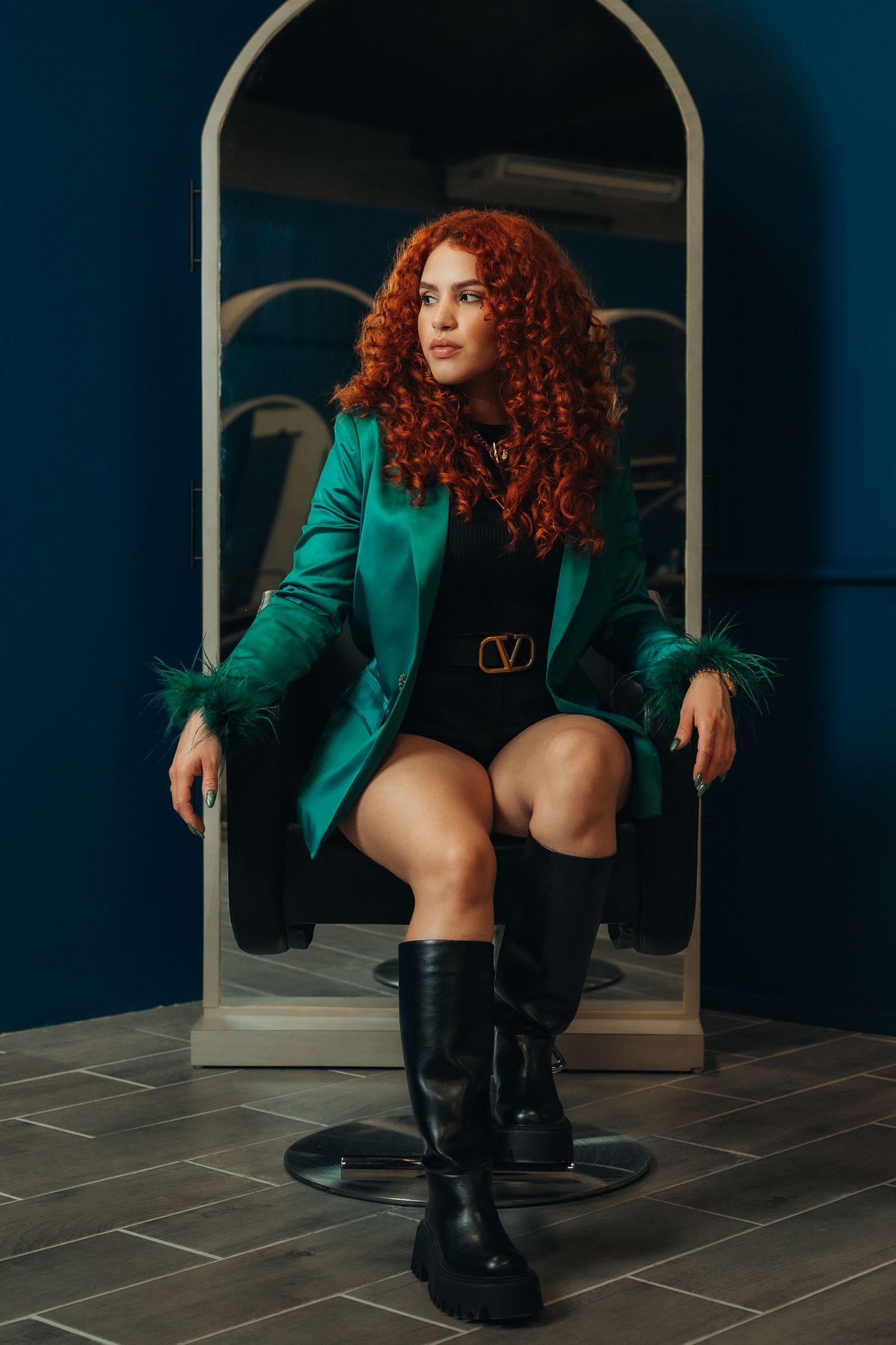 Woman with red curly hair in green Valentino blazer and black boots seated in chair against blue wall.