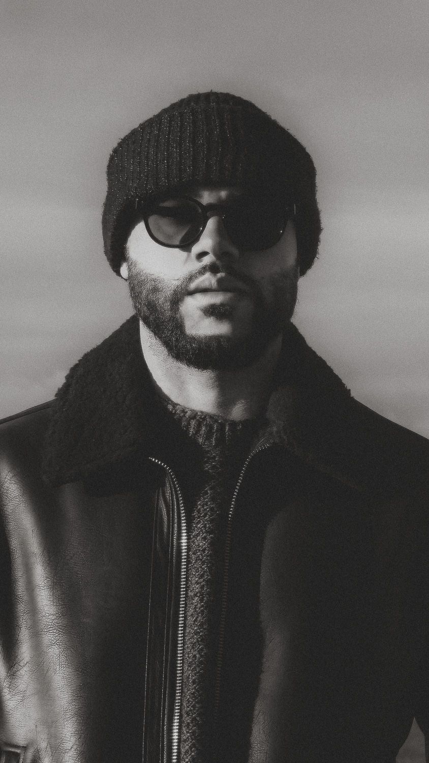 Black and white portrait of a bearded man wearing a knit beanie, sunglasses, and a leather jacket with shearling collar.