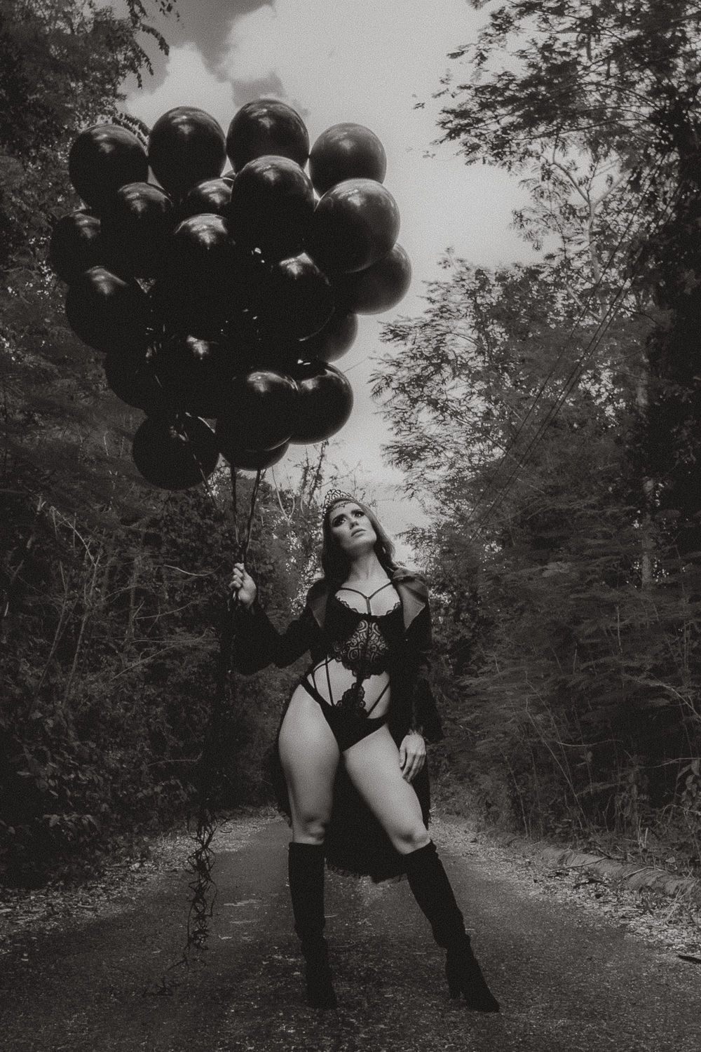 Black and white birthday portrait of a woman holding dark balloons while posing in a wooded outdoor setting
