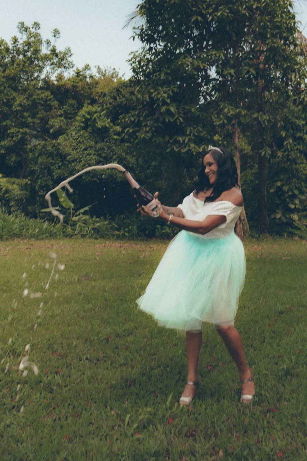 Woman in a white and mint dress popping a champagne bottle outdoors during a celebratory birthday photoshoot