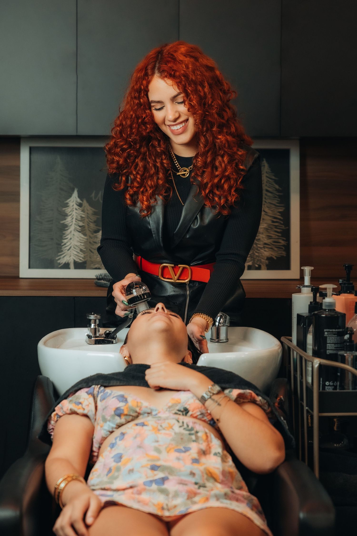 A smiling redhead hairstylist washes a client's hair at a salon shampoo bowl in a modern barbershop.