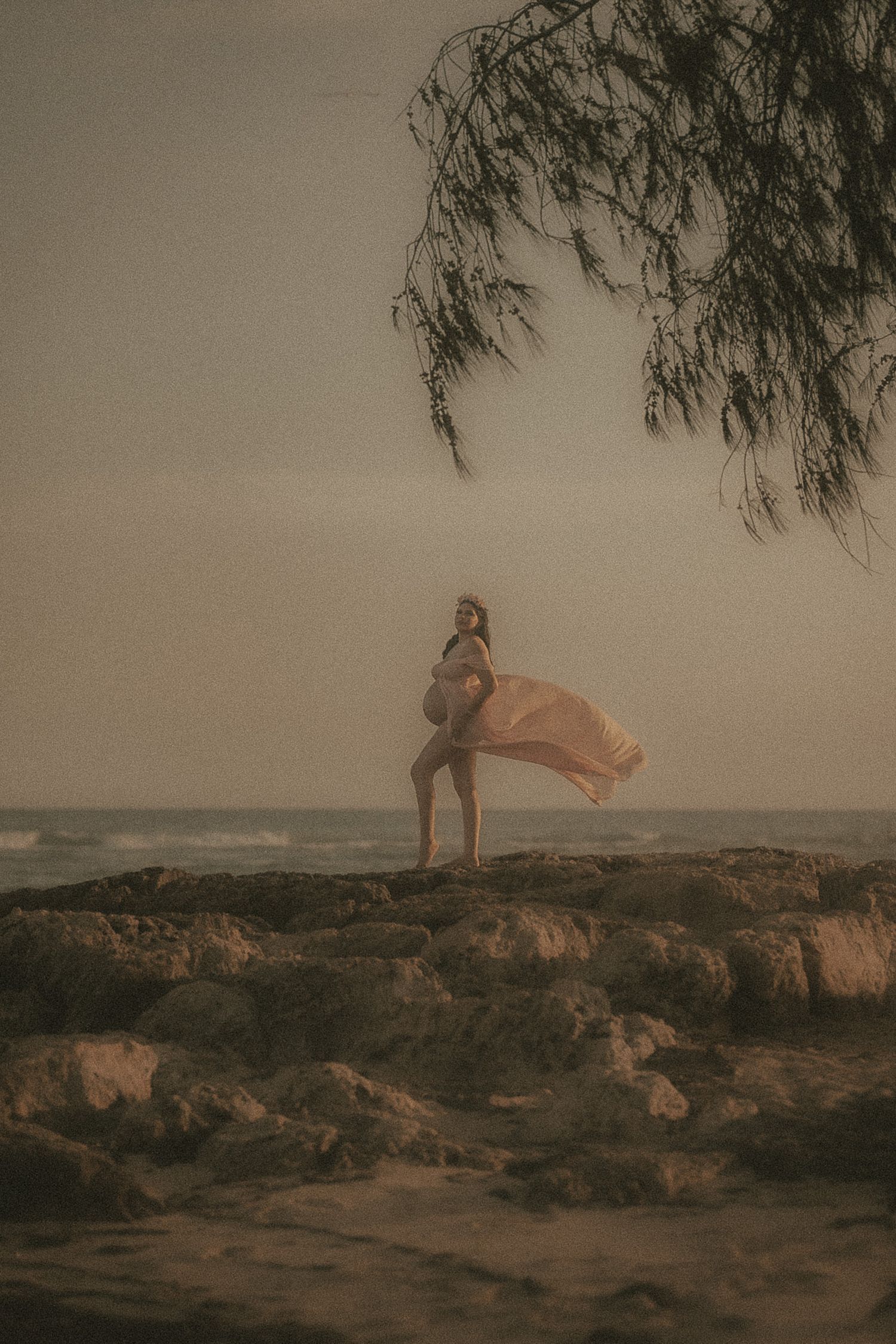 Expecting mother posing on rocky coastline with flowing maternity fabric at sunset
