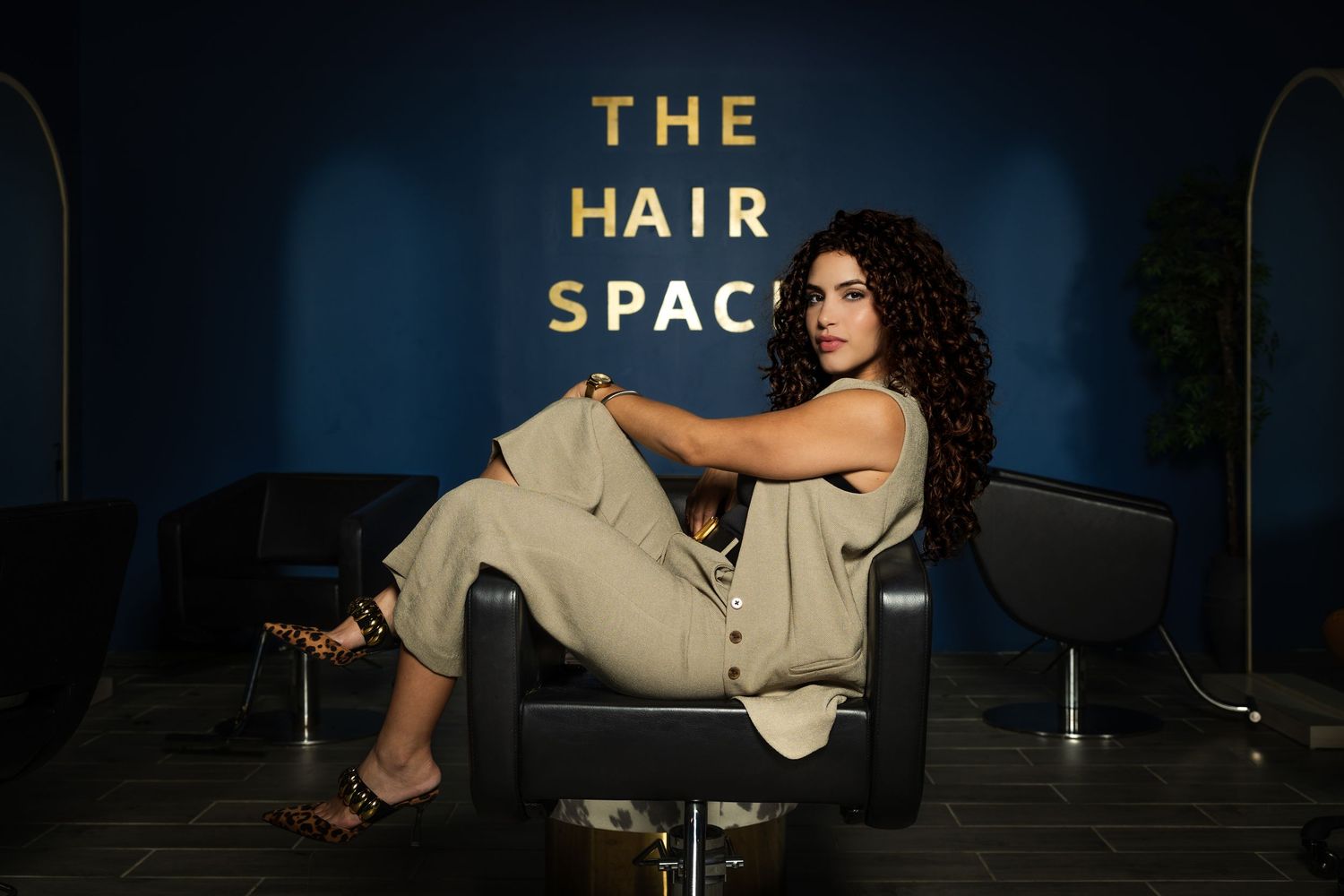 Woman in beige jumpsuit poses in salon chair at a modern, blue-walled hair salon.