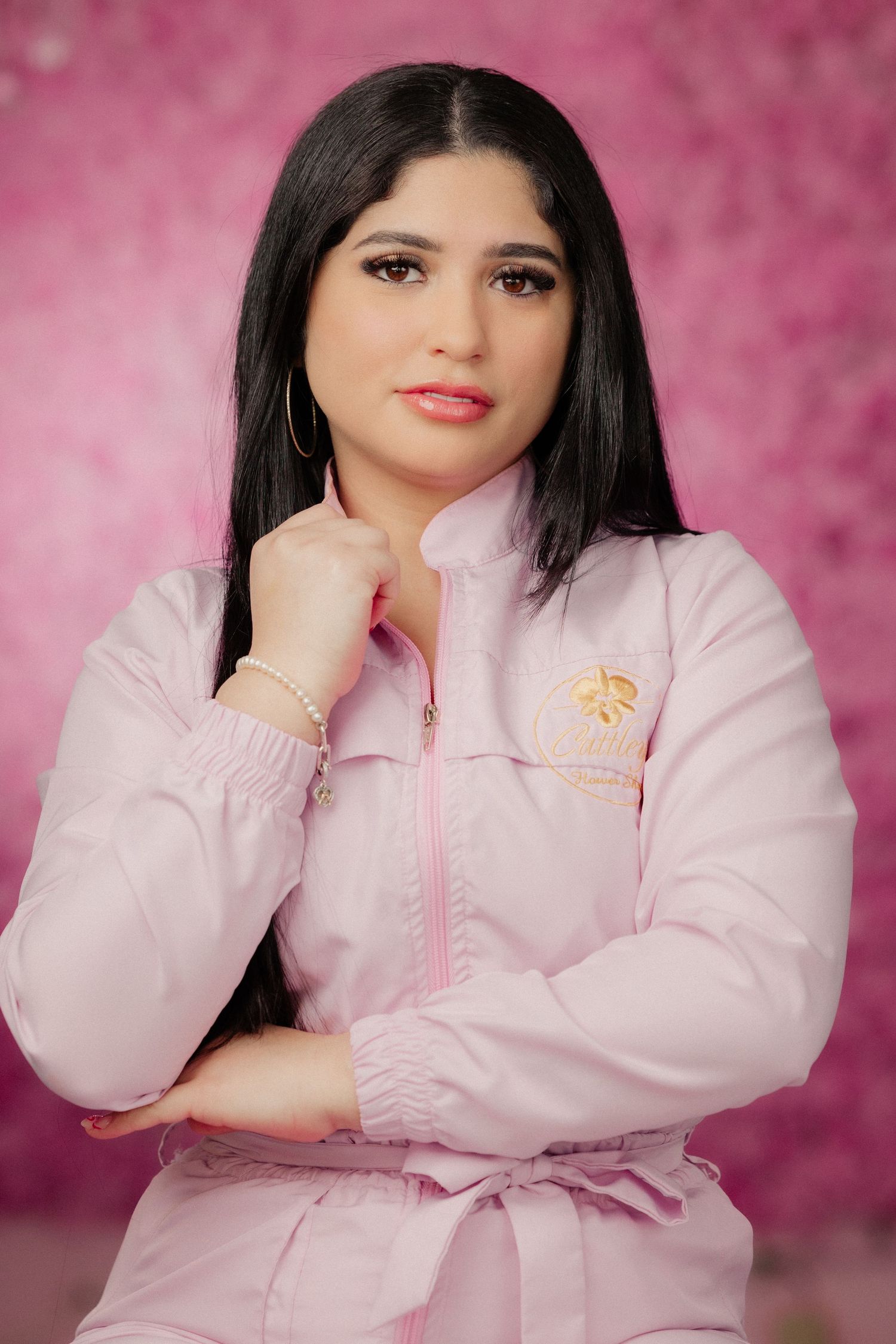 A young woman in a pink zip-up jacket poses confidently against a pink floral background.