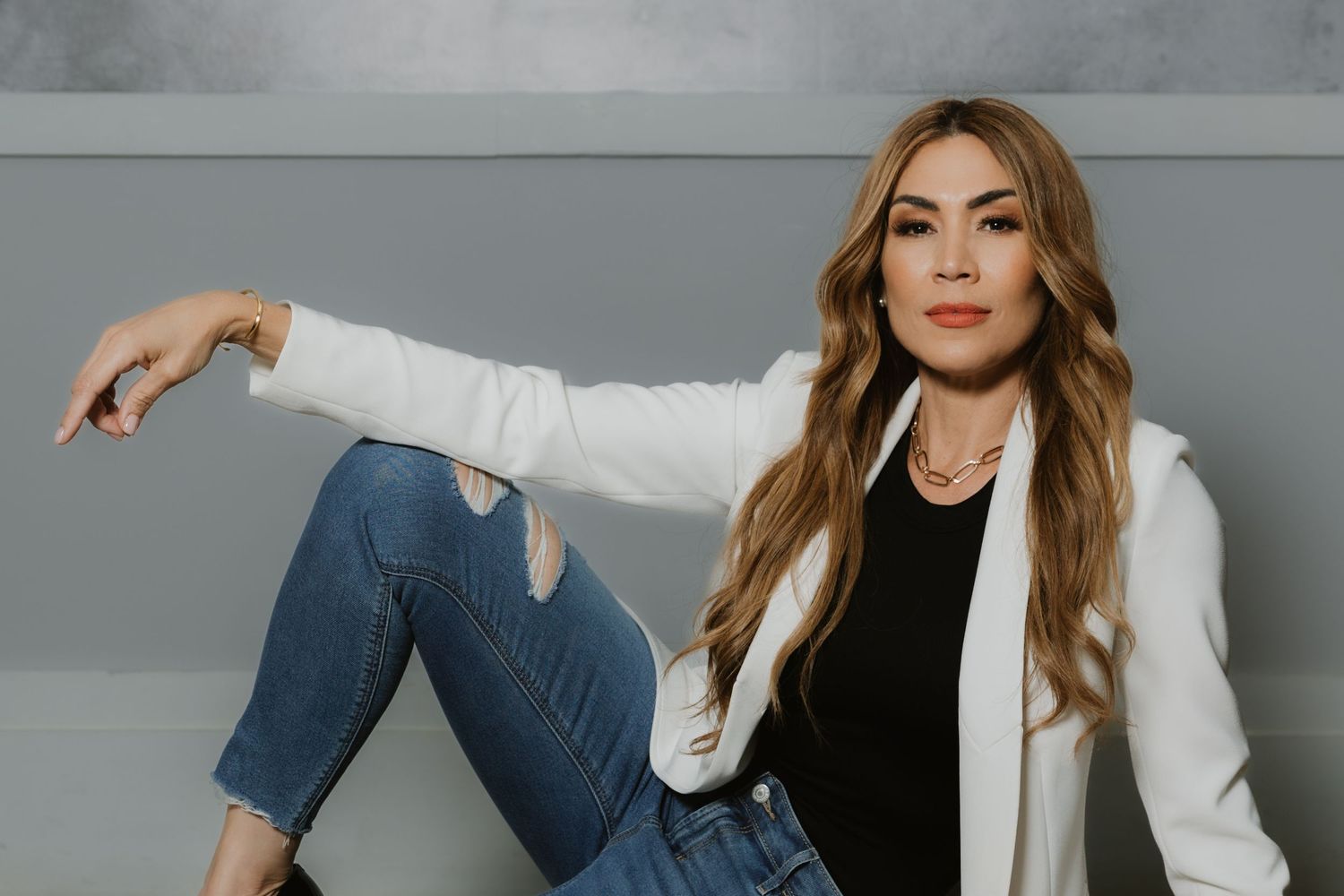 Woman in white blazer, black top, and ripped jeans sits confidently against a gray wall with long blonde hair.