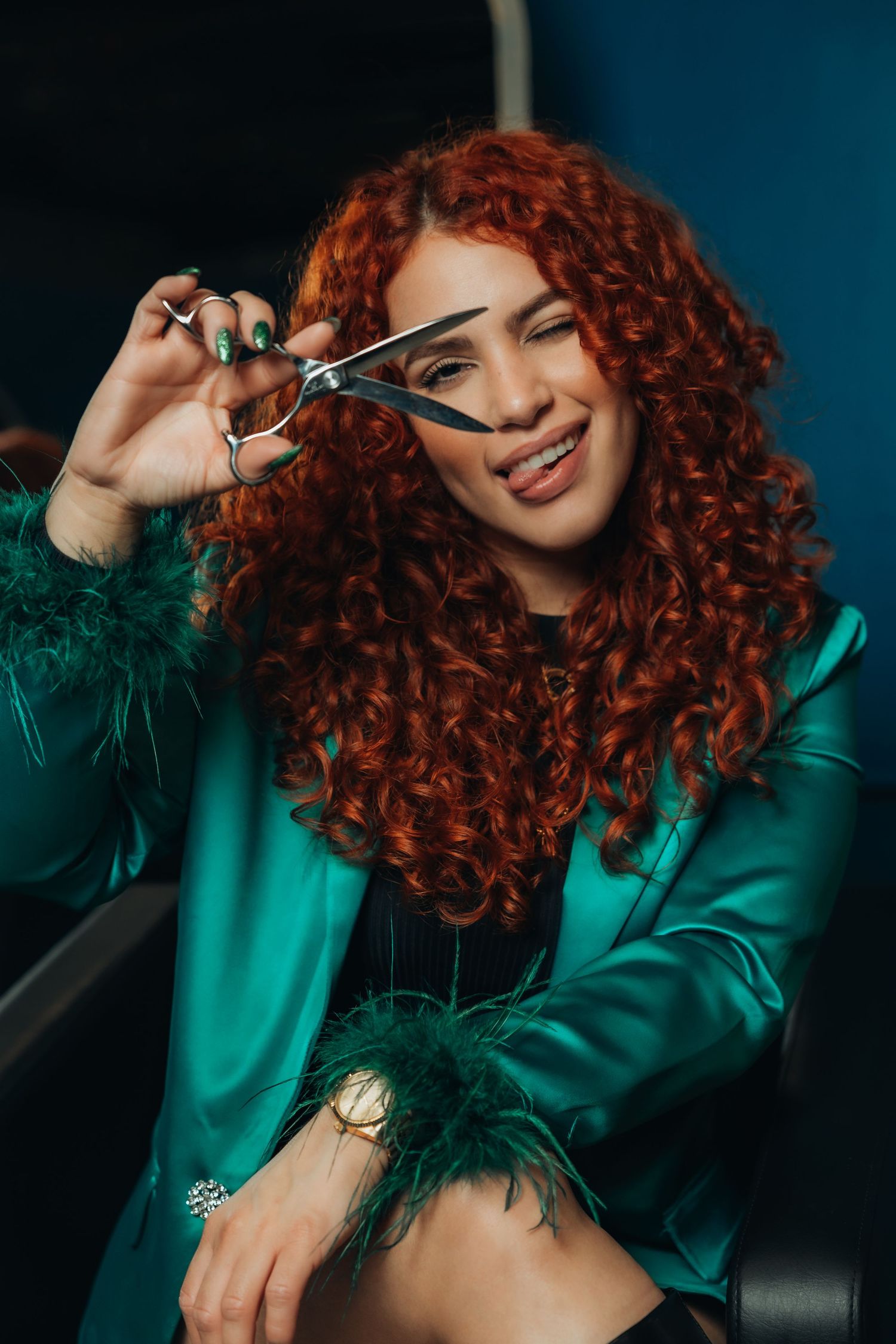 Red-haired woman in teal jacket holds scissors near face, smiling playfully in a salon setting.