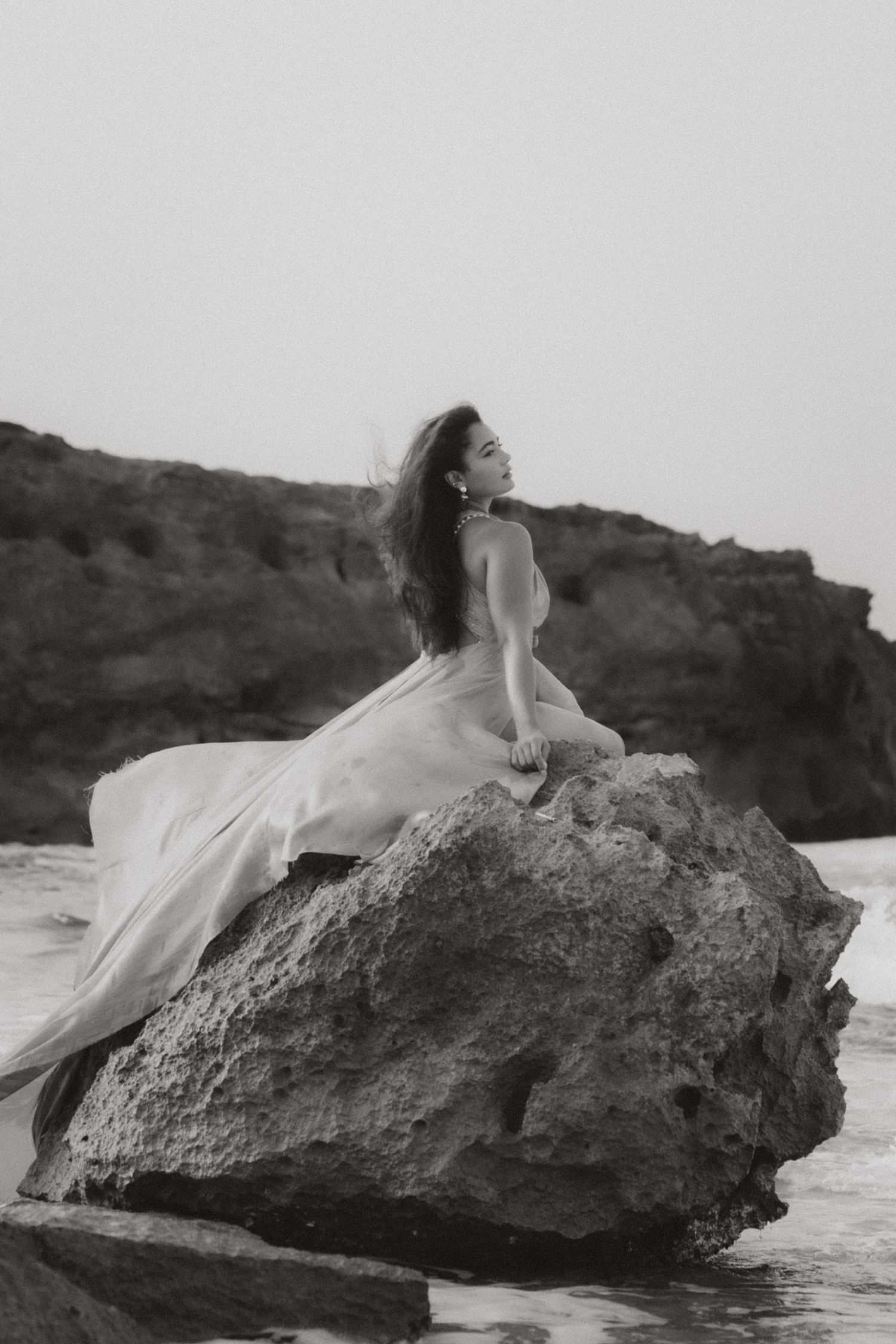 Black and white birthday portrait of a woman in a flowing white dress sitting on a rocky coastal landscape