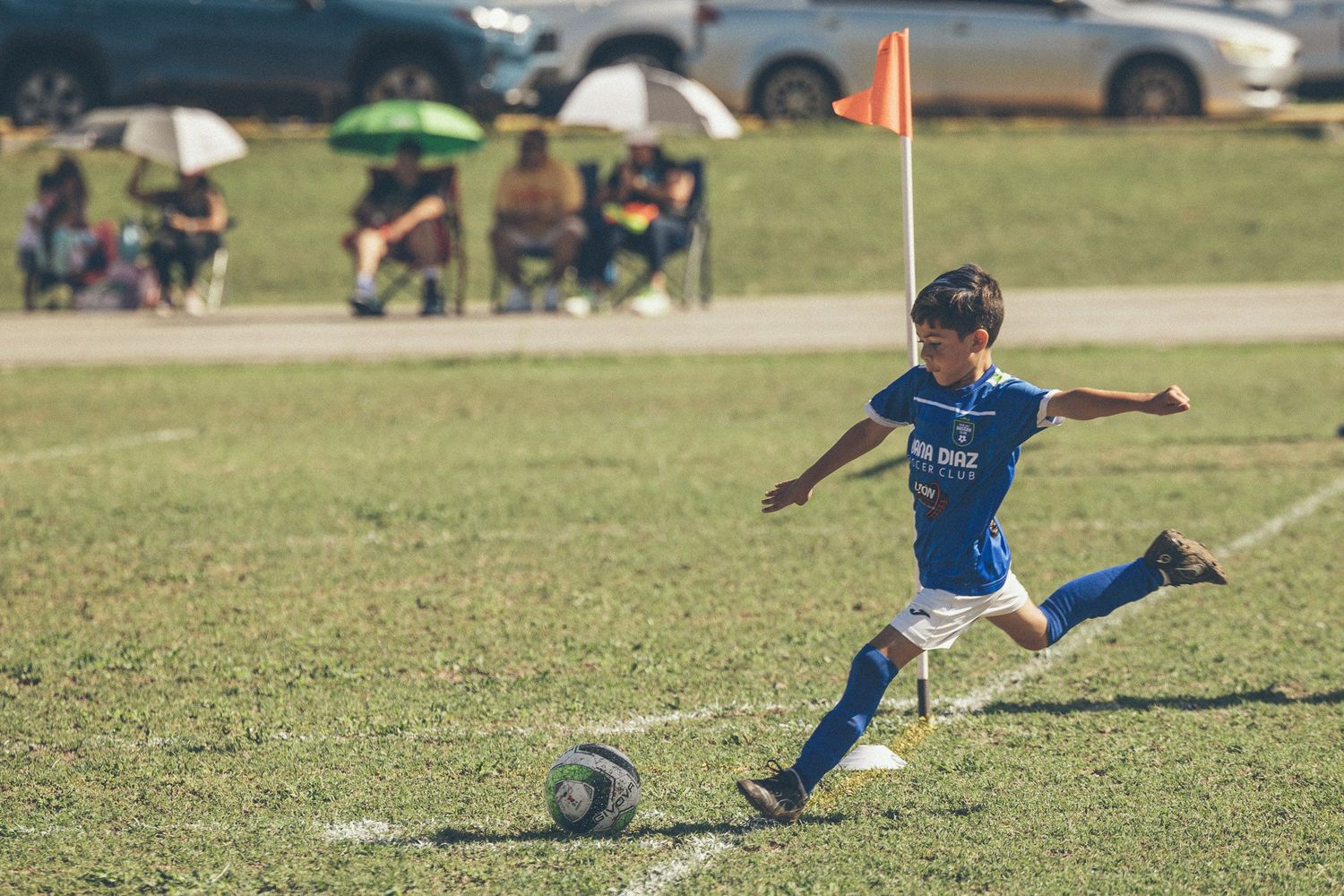 Young boy in blue uniform kicking soccer ball on green field during youth soccer game with spectators in background.