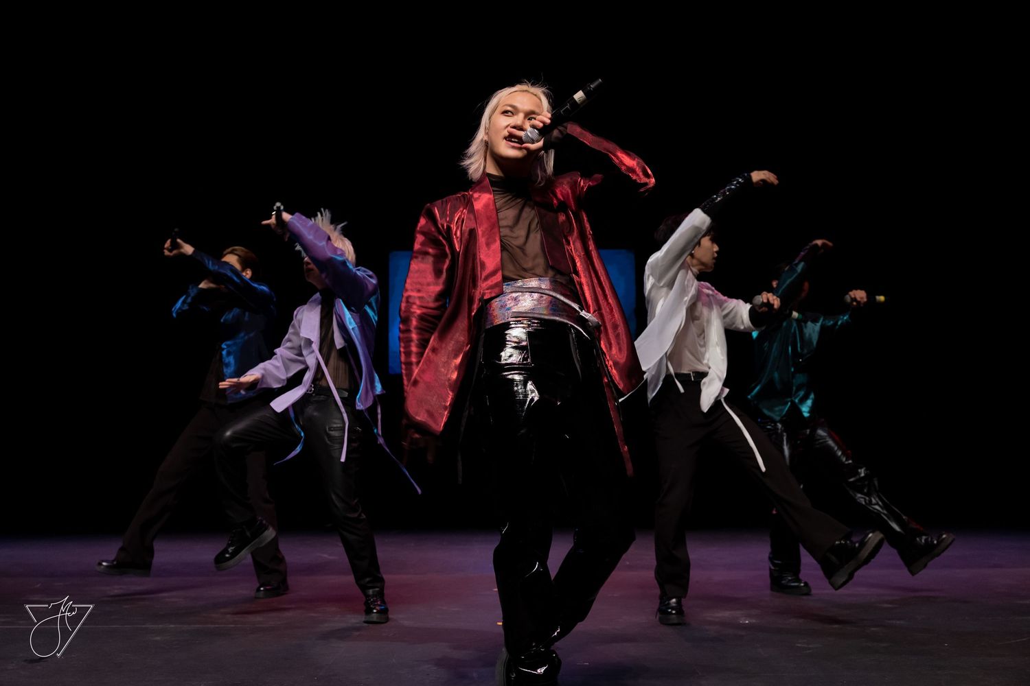 Performers in vibrant costumes dance on stage, led by a blonde dancer in a red jacket and black pants.