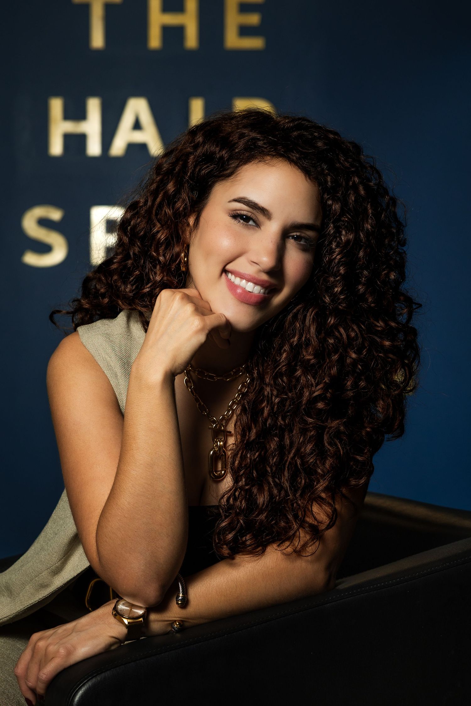 Smiling woman with long curly brown hair poses confidently at The Hair salon, wearing a green top and gold jewelry.