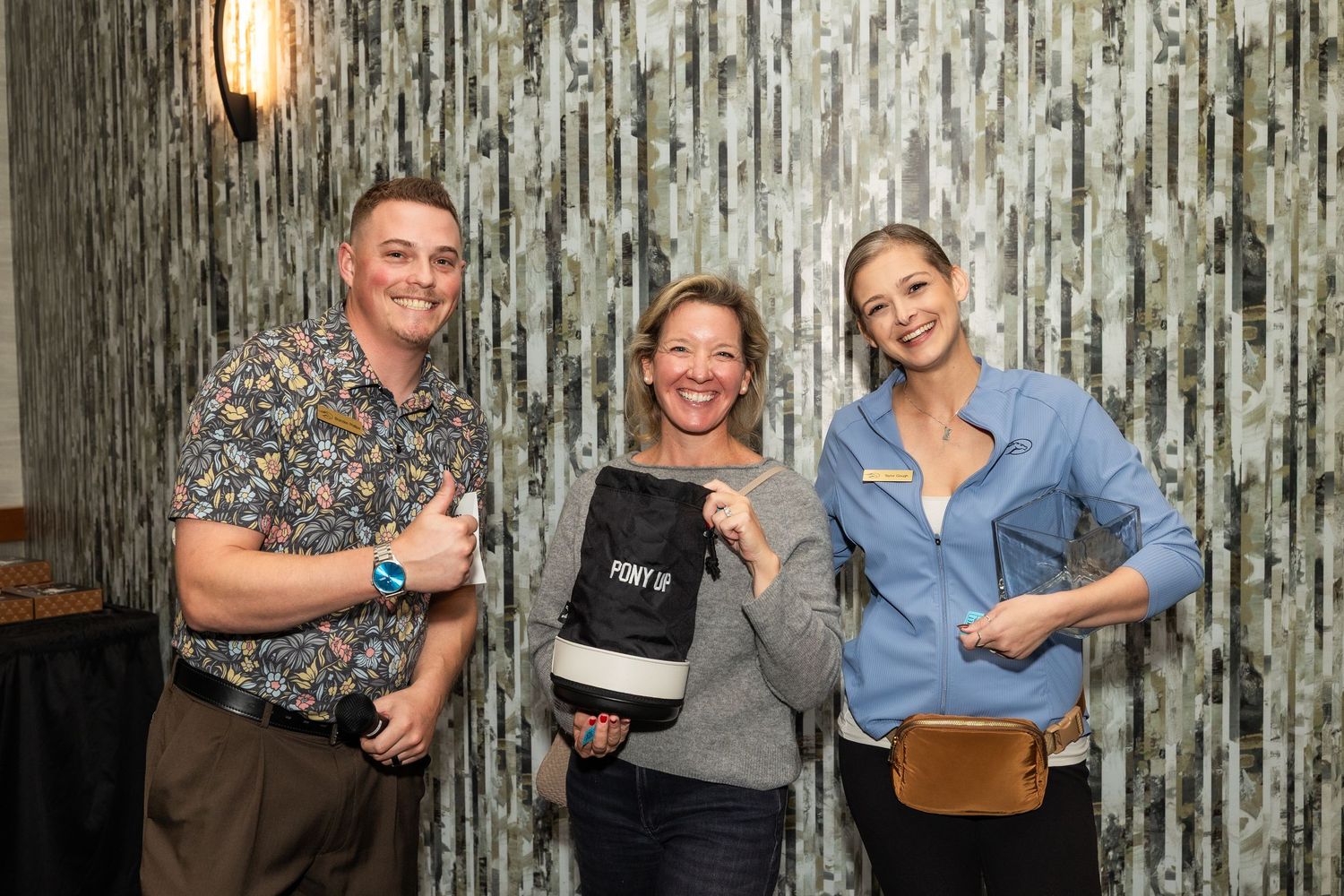 Three smiling people posing together holding a Pony Up branded bag at an indoor event.