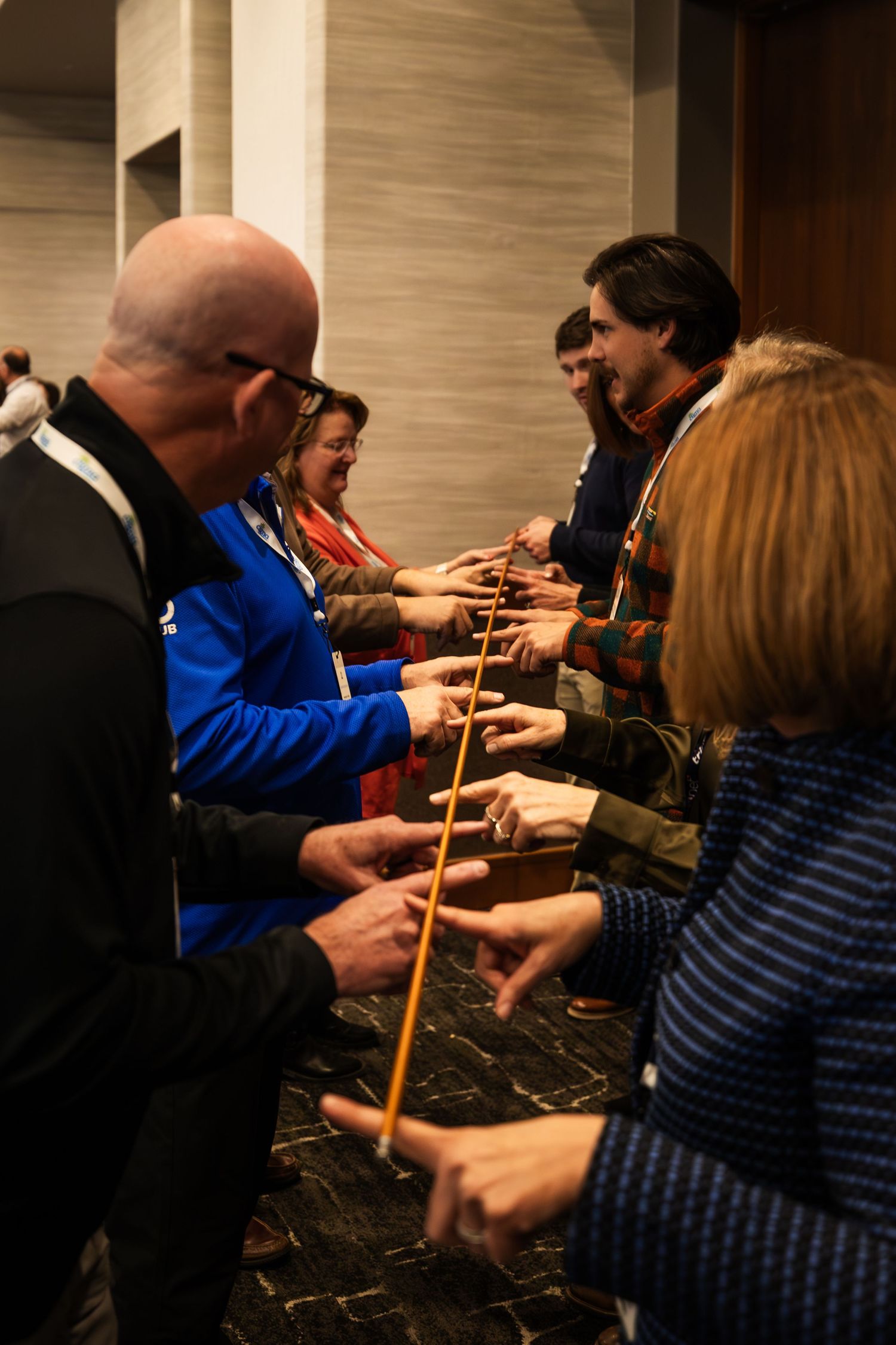 Group of people standing in two lines facing each other, passing a wooden stick in a team-building activity indoors.