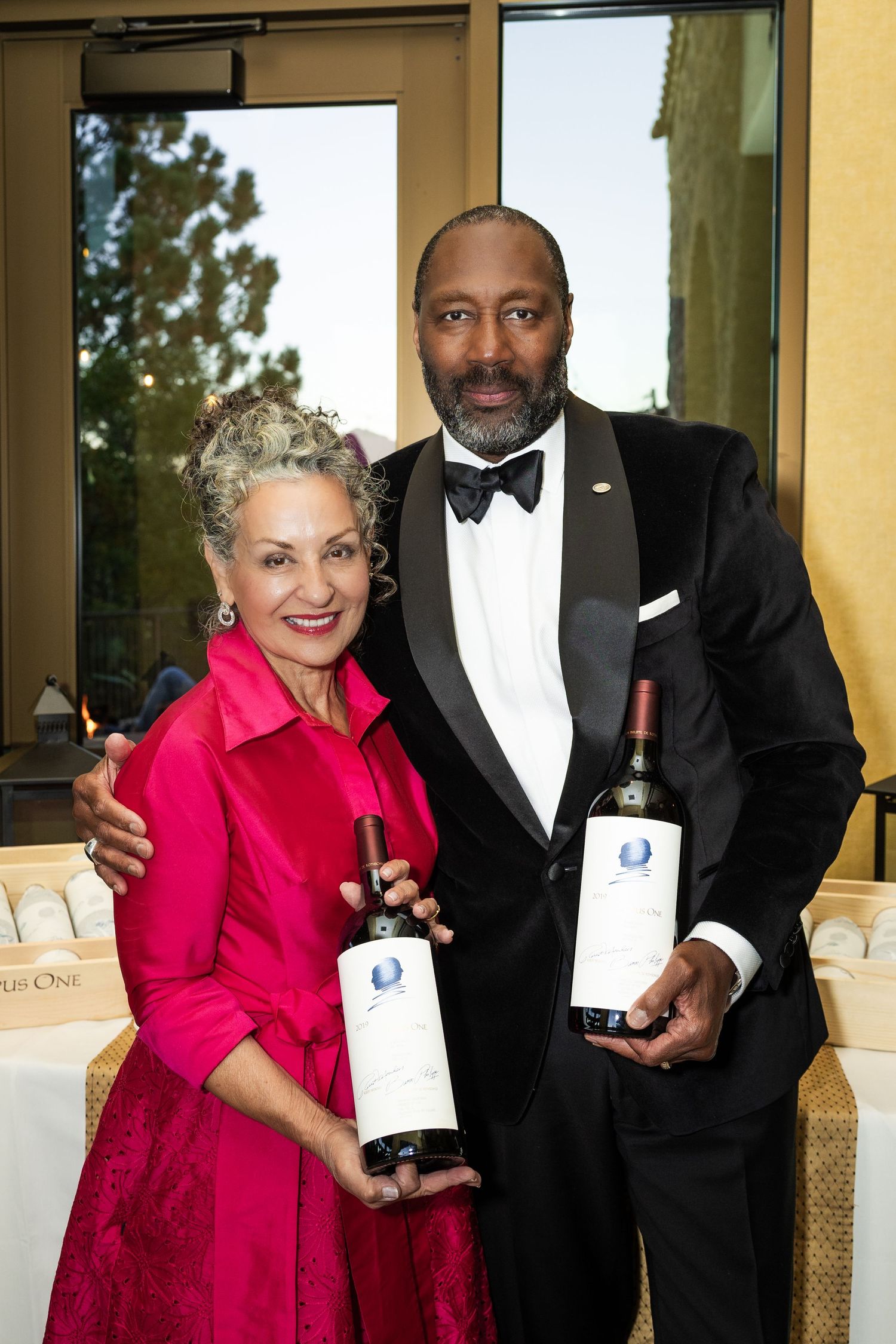 Elegantly dressed couple holding Opus One wine bottles at a formal event, woman in pink dress, man in tuxedo.
