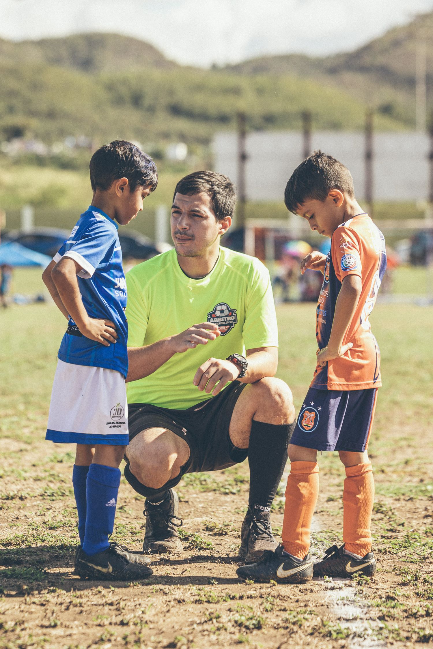 A soccer coach in a green jersey crouches down to talk to two young players in blue and orange uniforms on a field.