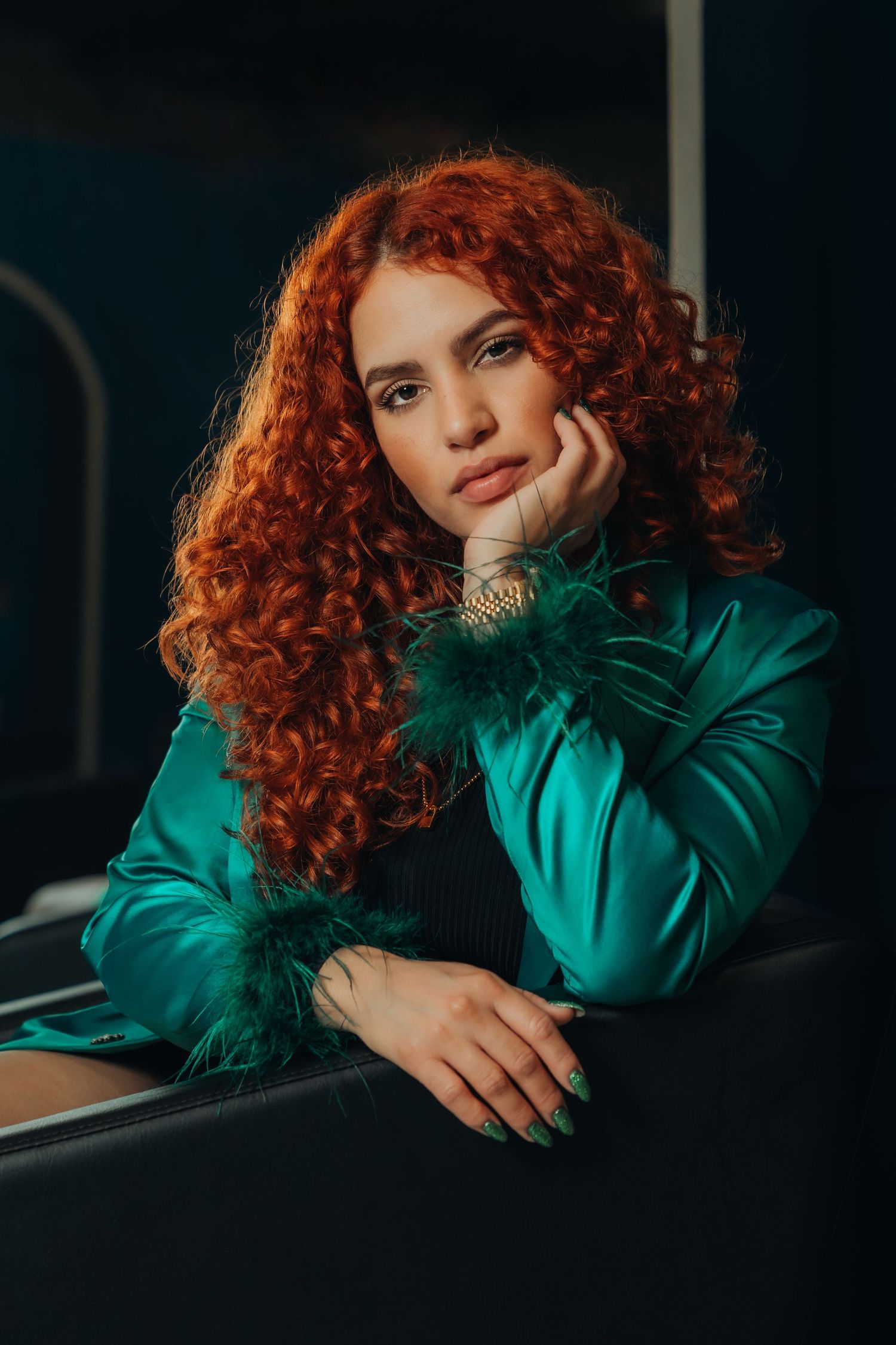 Young woman with curly red hair wearing a teal satin jacket with feather cuffs, resting her chin on her hand.