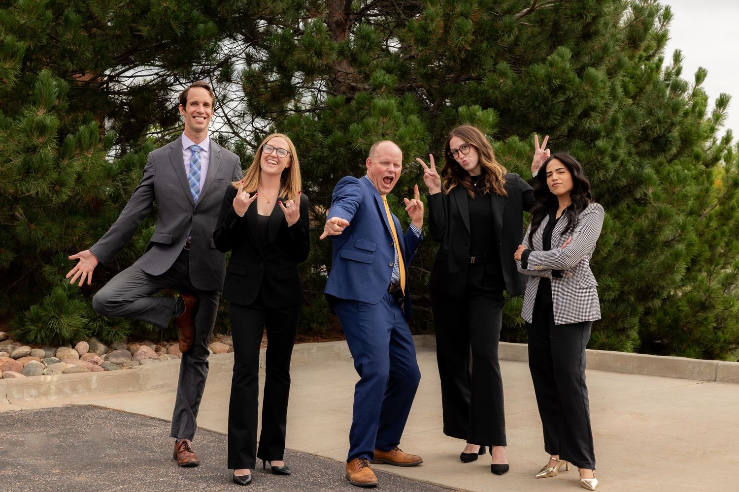 Five business professionals posing playfully outdoors in formal attire, smiling and making fun gestures together.
