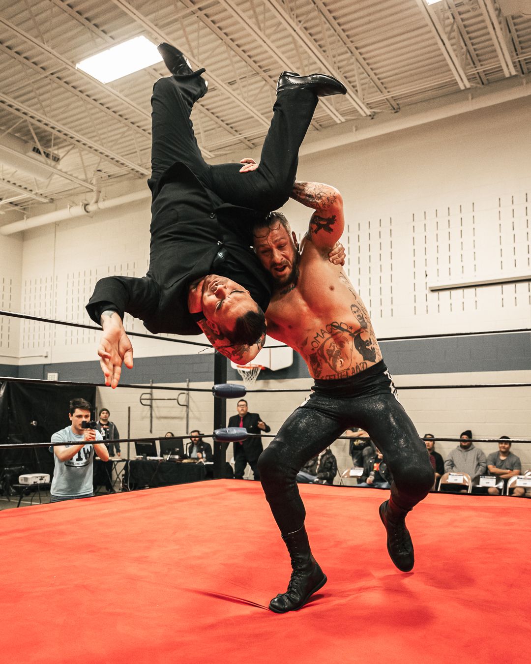 Two wrestlers perform a dramatic suplex move on a red ring mat during an indoor professional wrestling event.