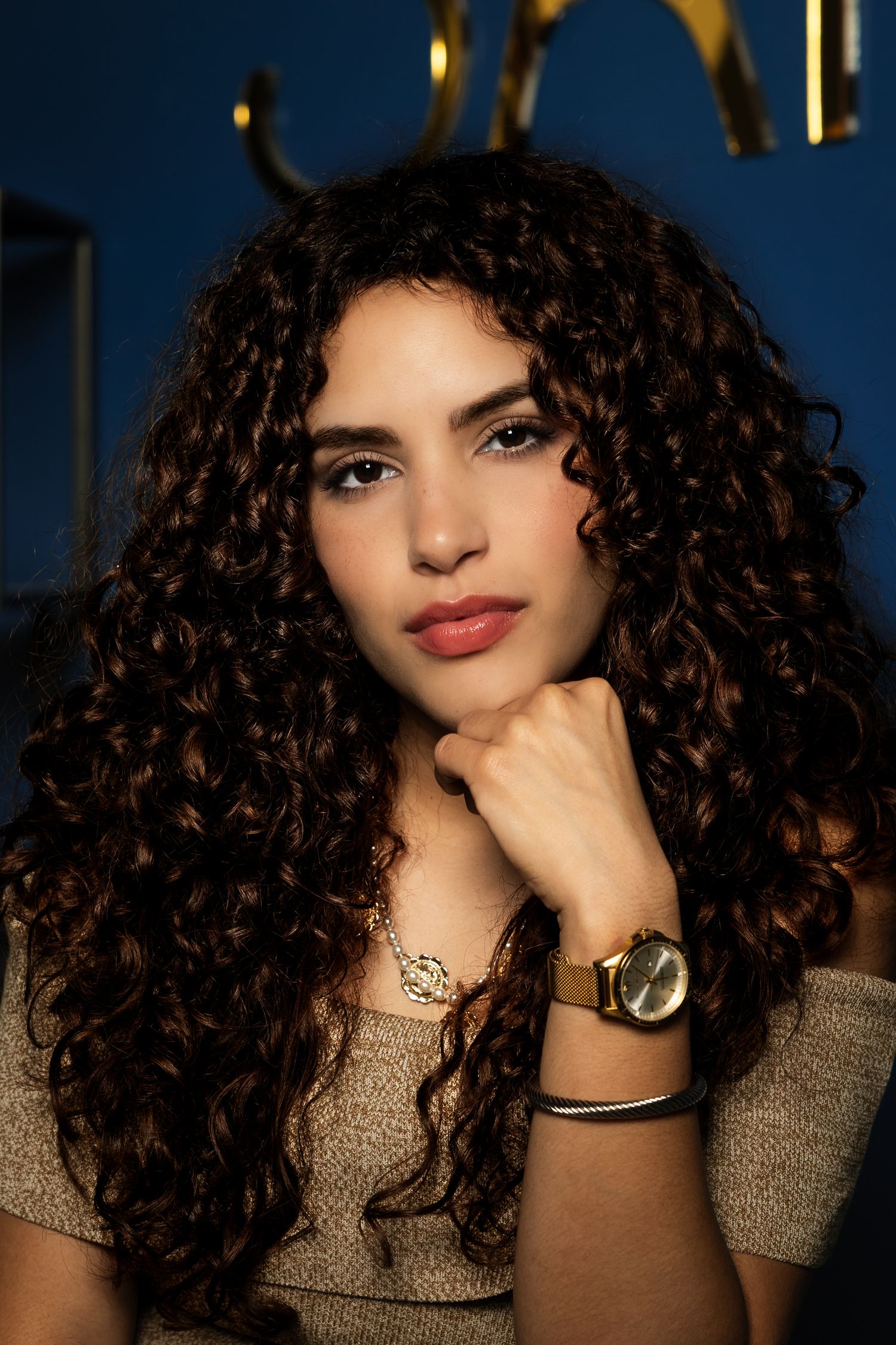 Young woman with long curly brown hair wearing a gold watch and necklace, posing against a blue background.