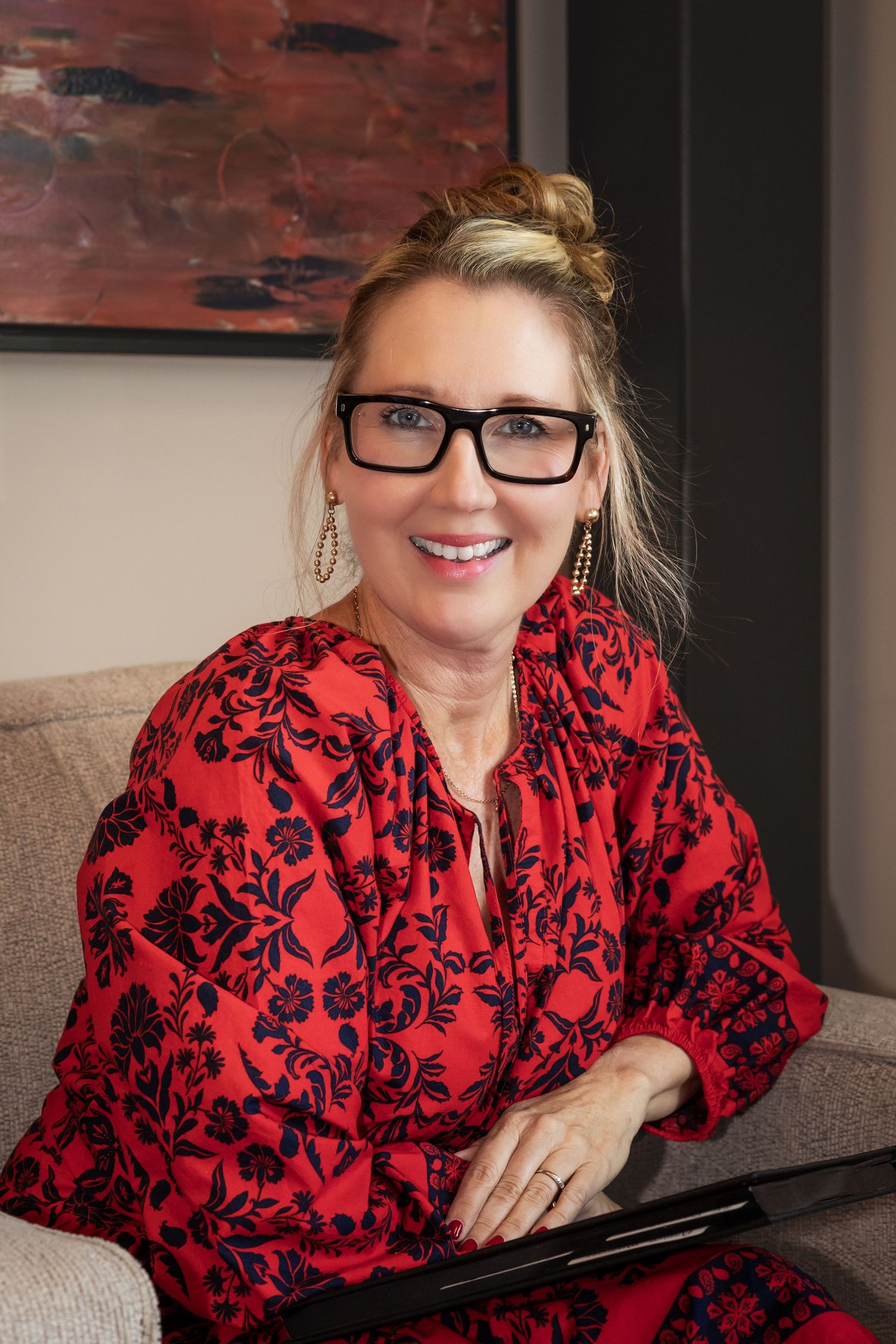 Smiling woman in red floral blouse with black glasses, sitting on sofa holding a tablet in a modern interior.