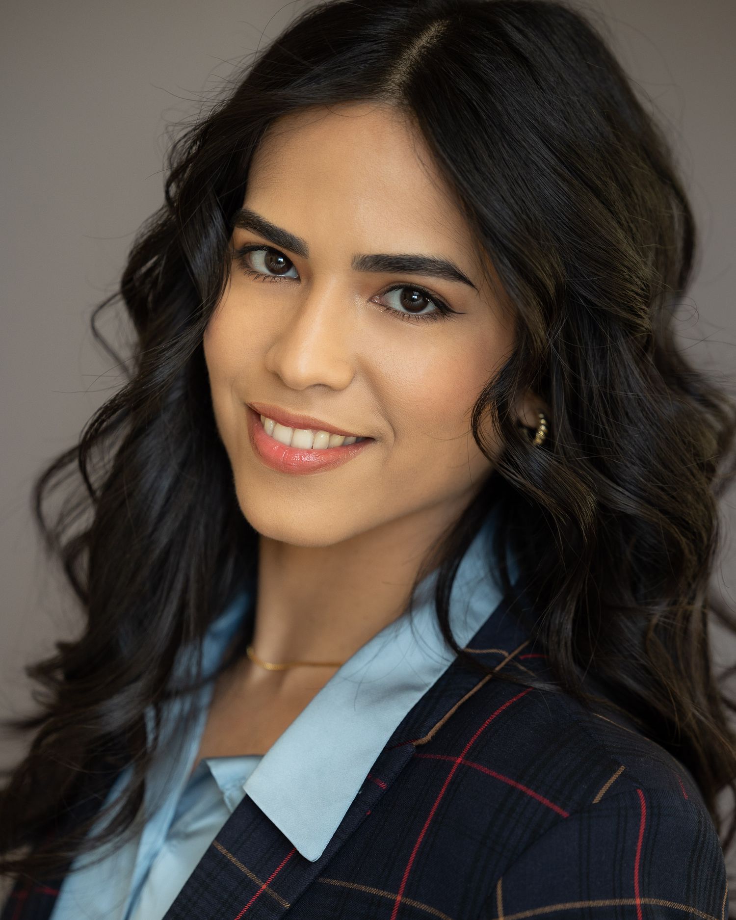 Professional headshot of a young woman with long dark wavy hair wearing a plaid blazer and light blue shirt, smiling confidently.