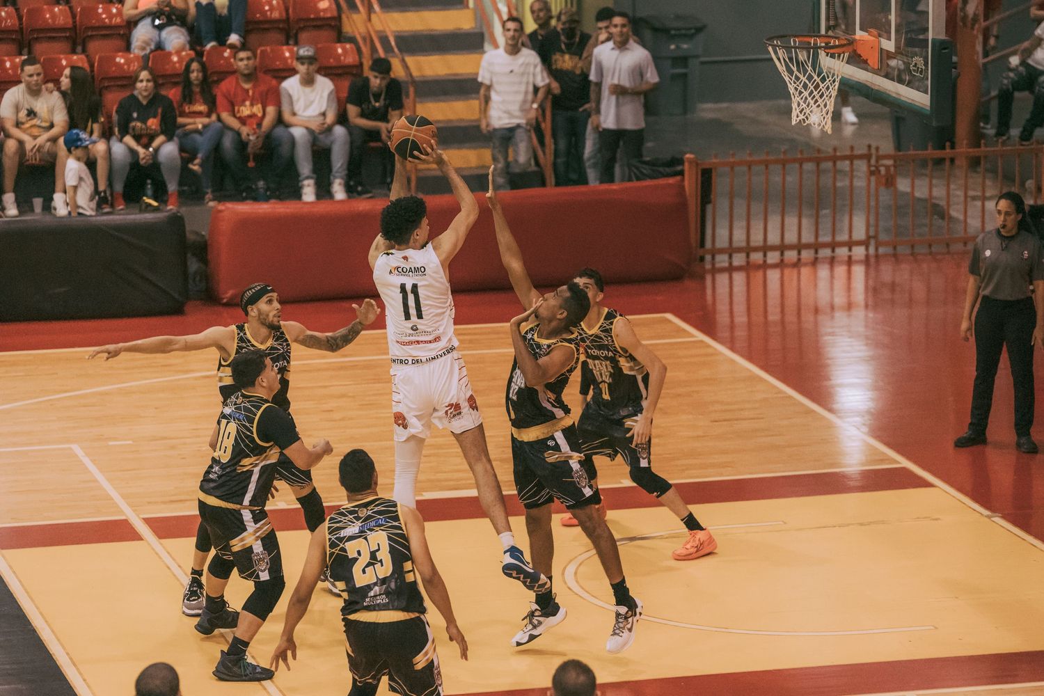 Basketball player #11 shoots over 5 defenders during an indoor game with fans watching in the stands.