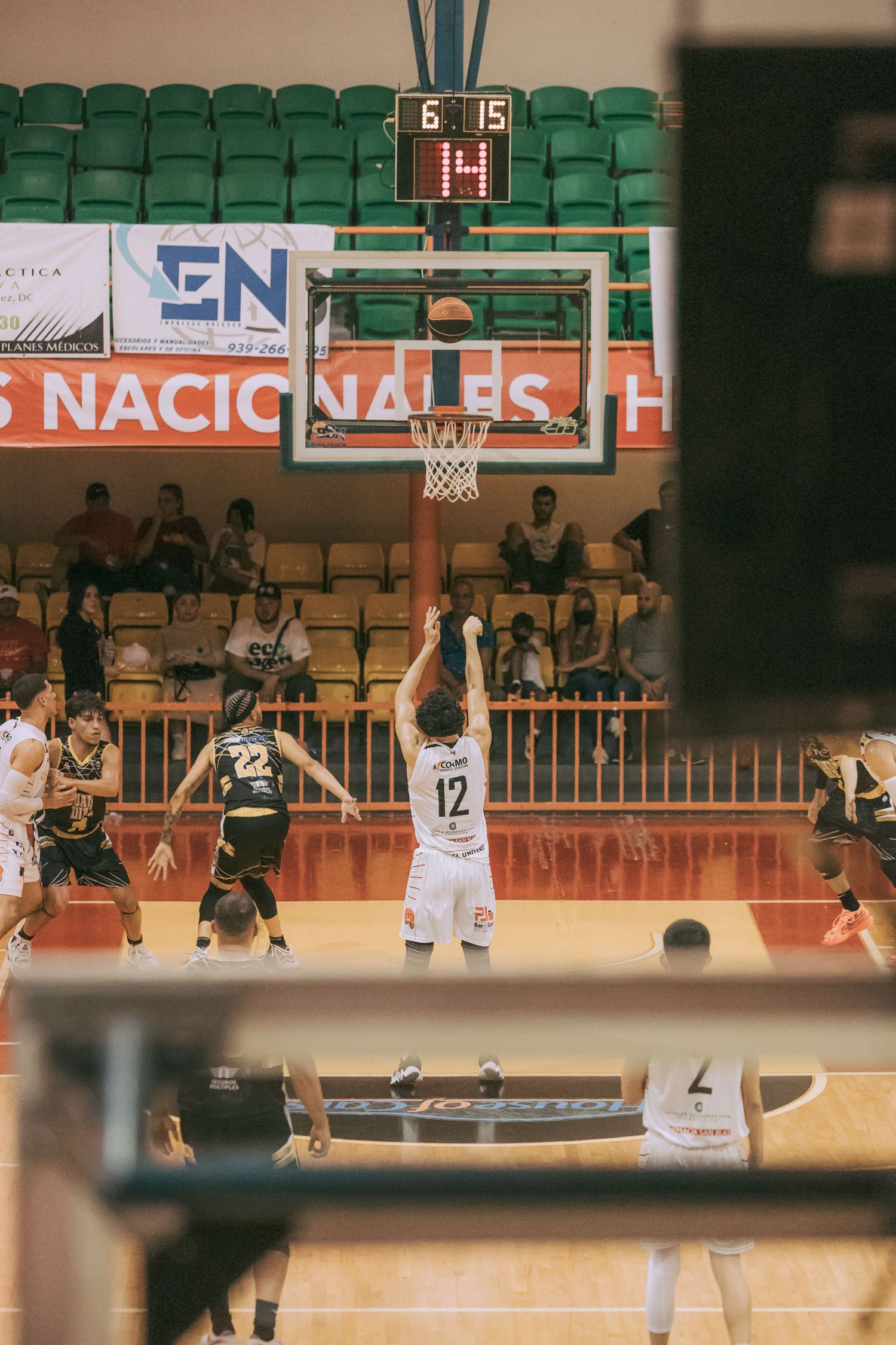 Player #12 attempts a free throw during an indoor basketball game with spectators watching in the stands.