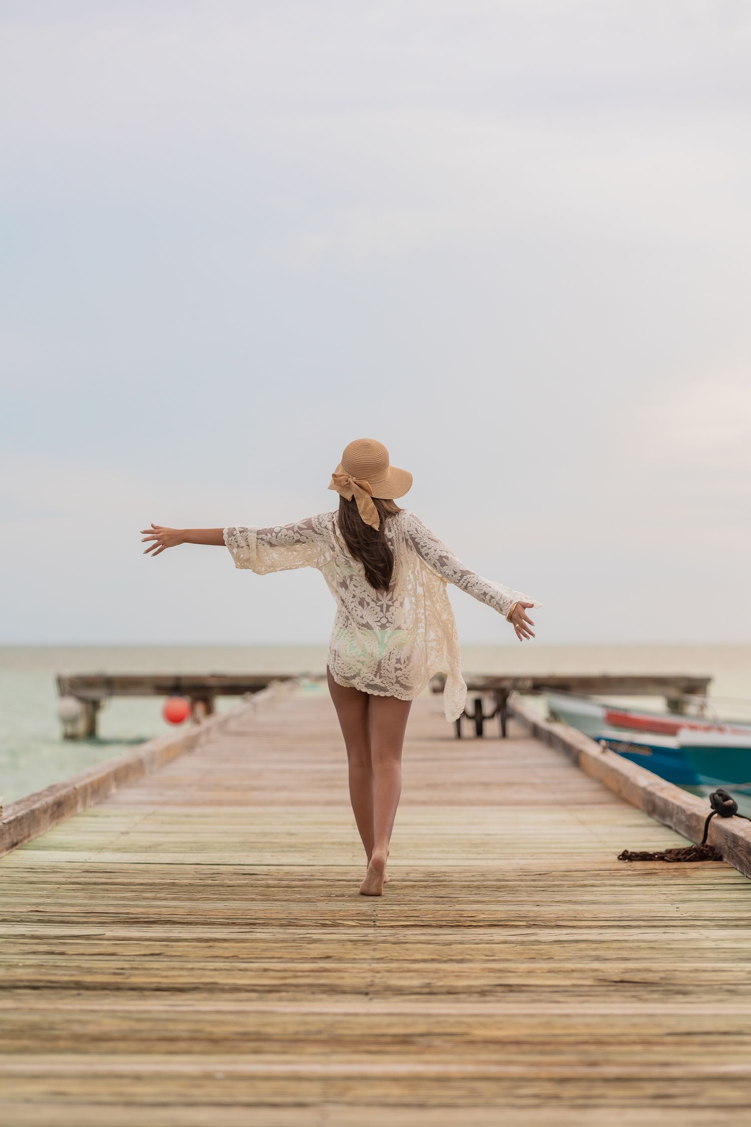 Lifestyle birthday portrait of a teenage woman in a sun hat walking along a seaside pier with her arms open