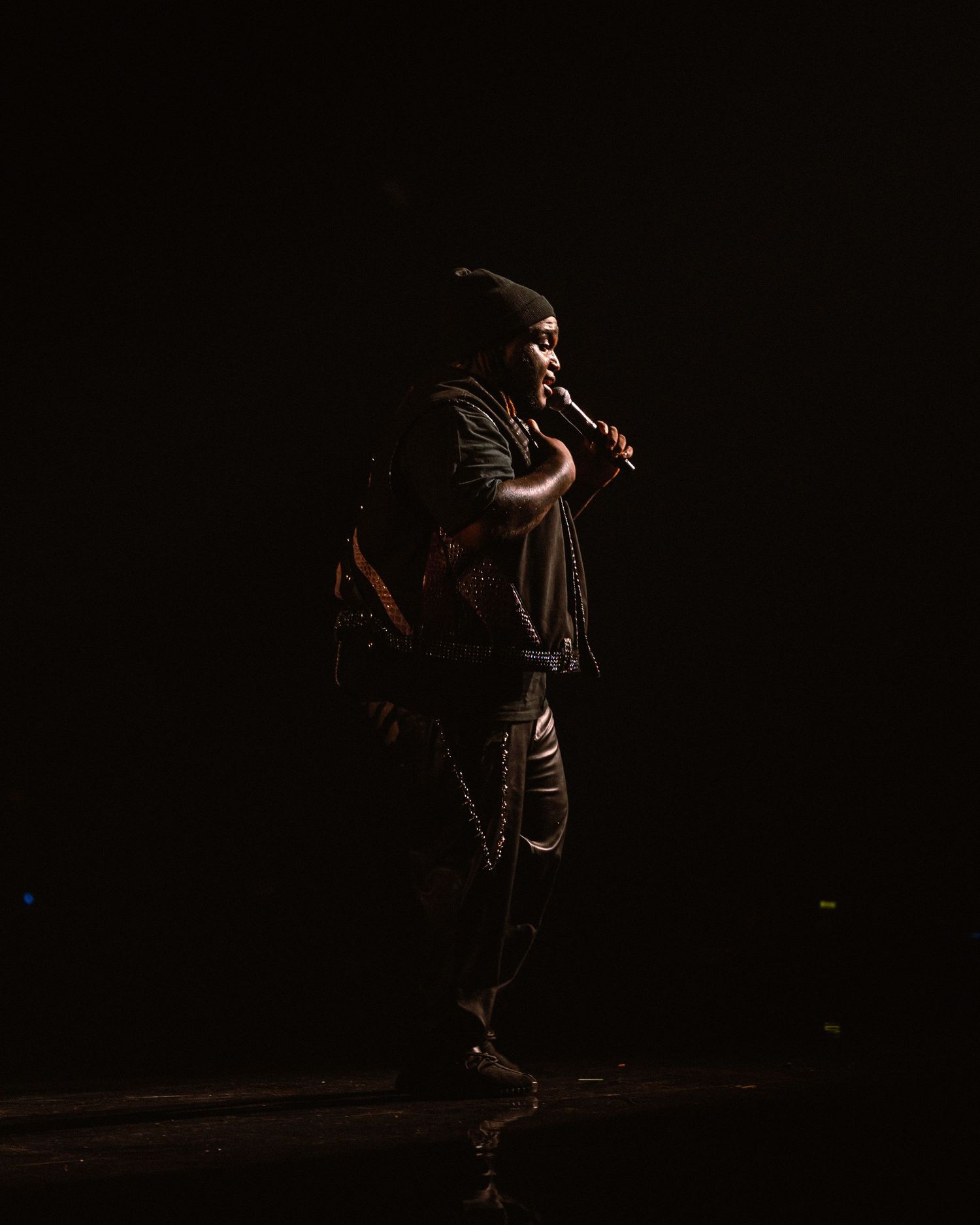 Performer on dark stage holding microphone, dramatically lit from above during live concert performance.