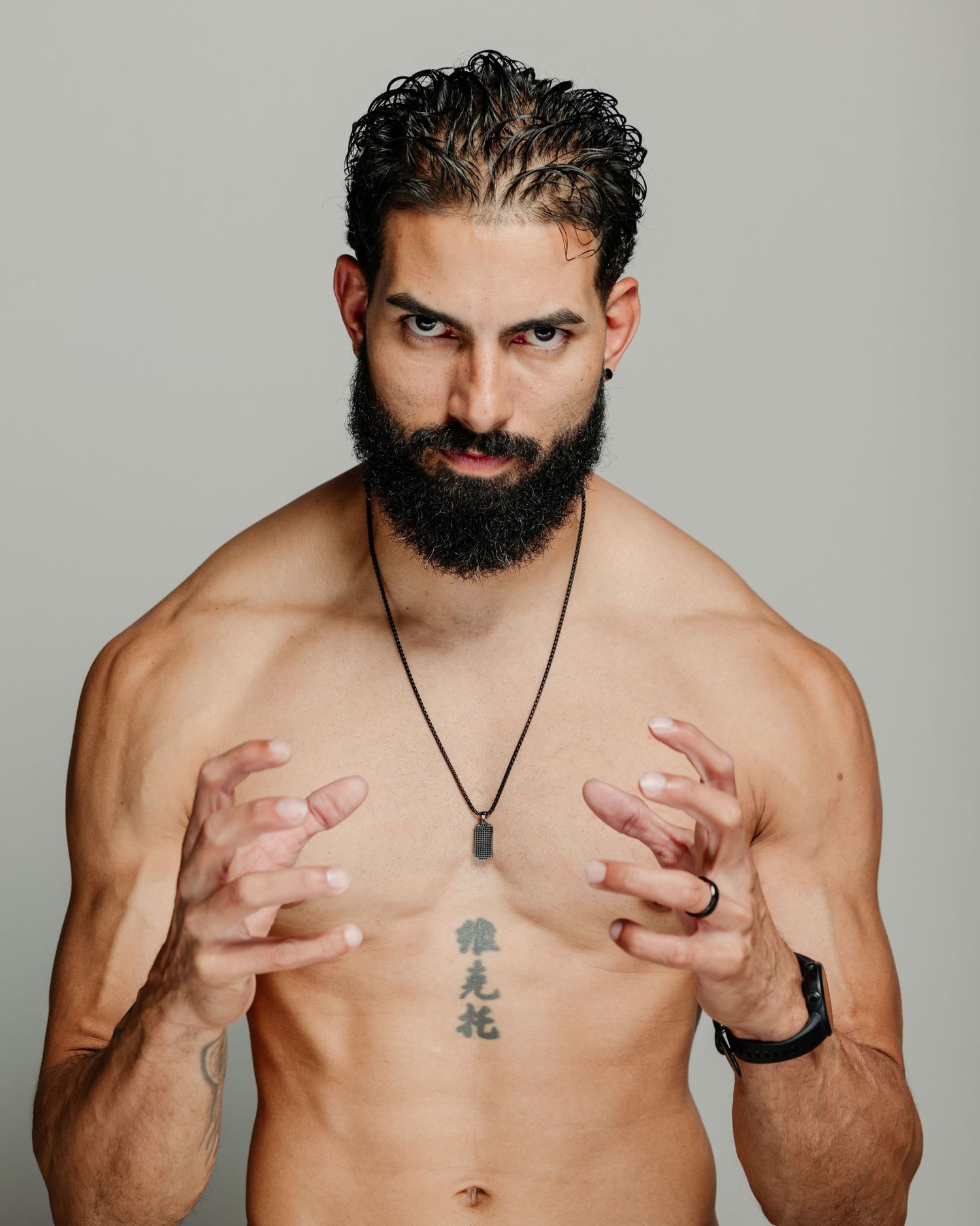 Muscular bearded man with chest tattoos and necklace posing shirtless against gray background.