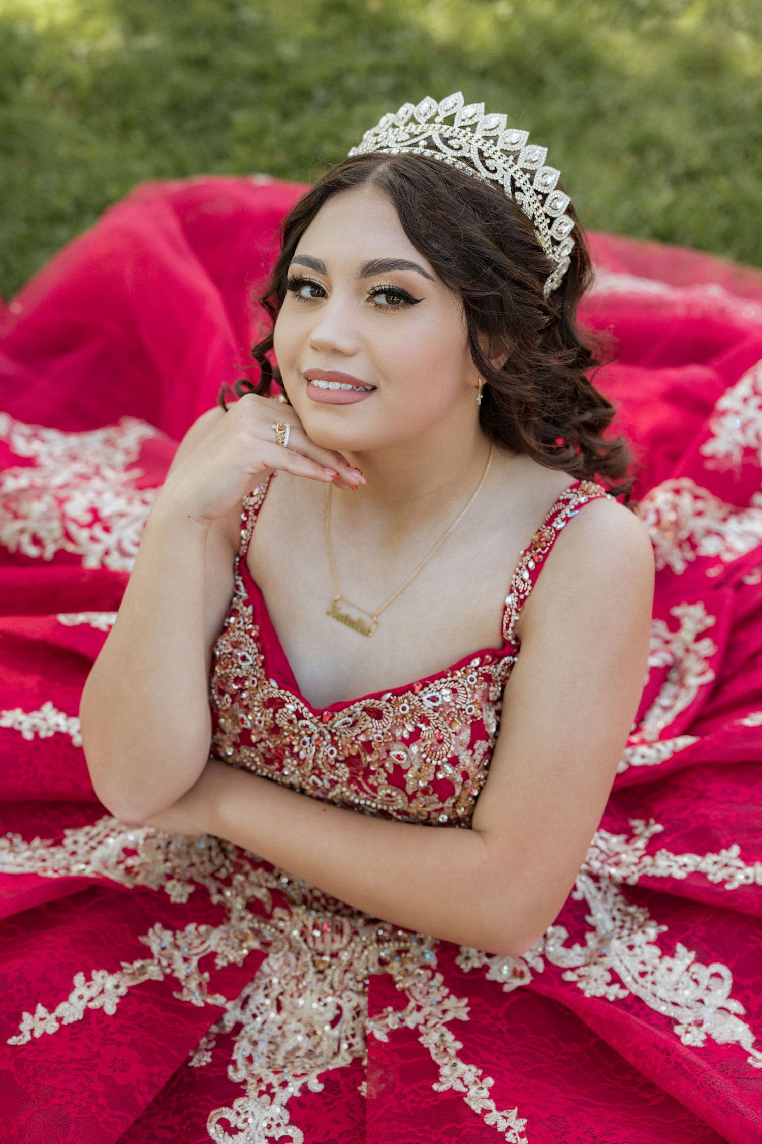 Young woman in red quinceañera gown with gold lace, crystal tiara, and gold necklace poses gracefully outdoors.