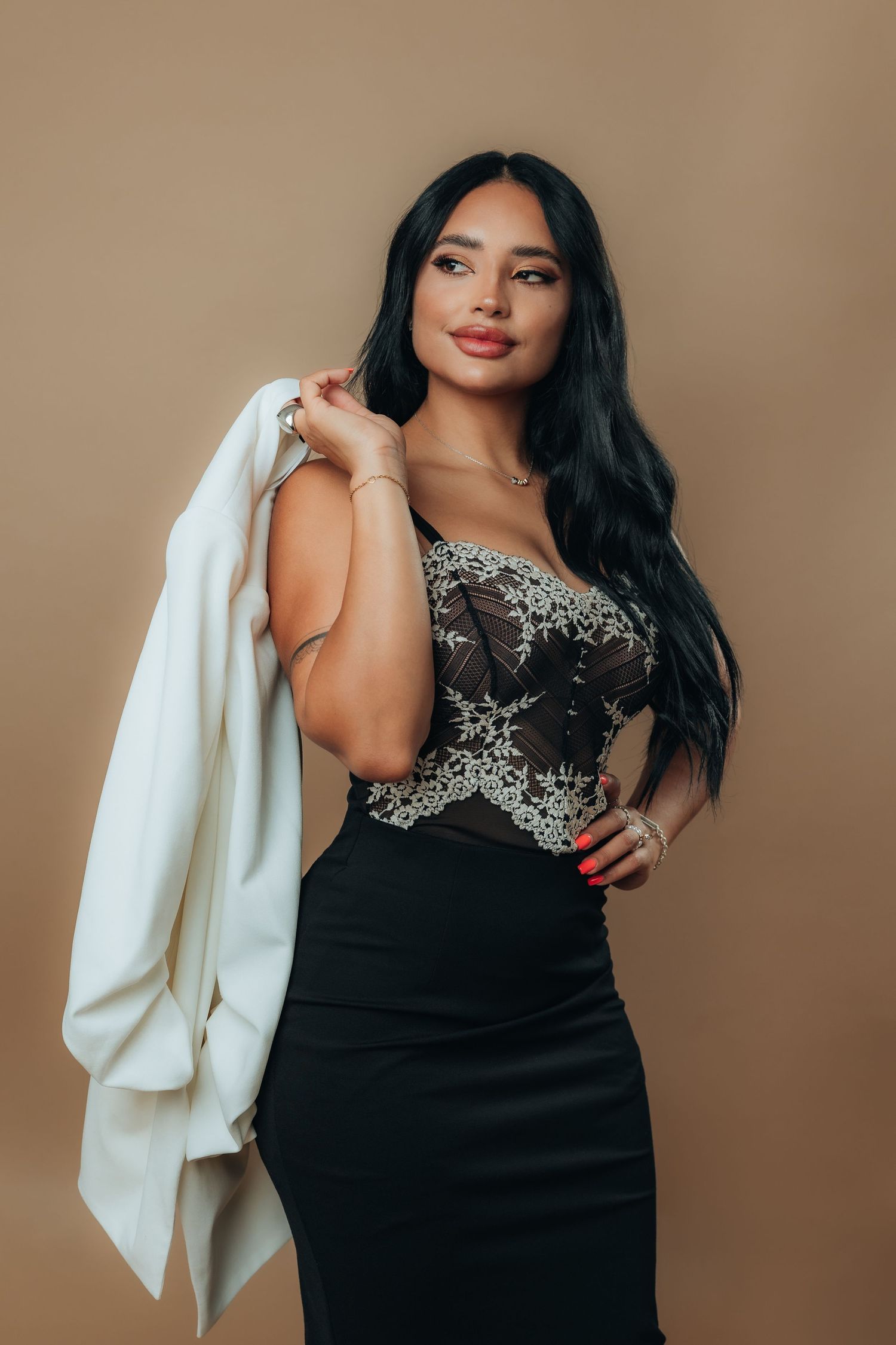 Woman in black lace corset gown holding white blazer over shoulder, posing against beige background.