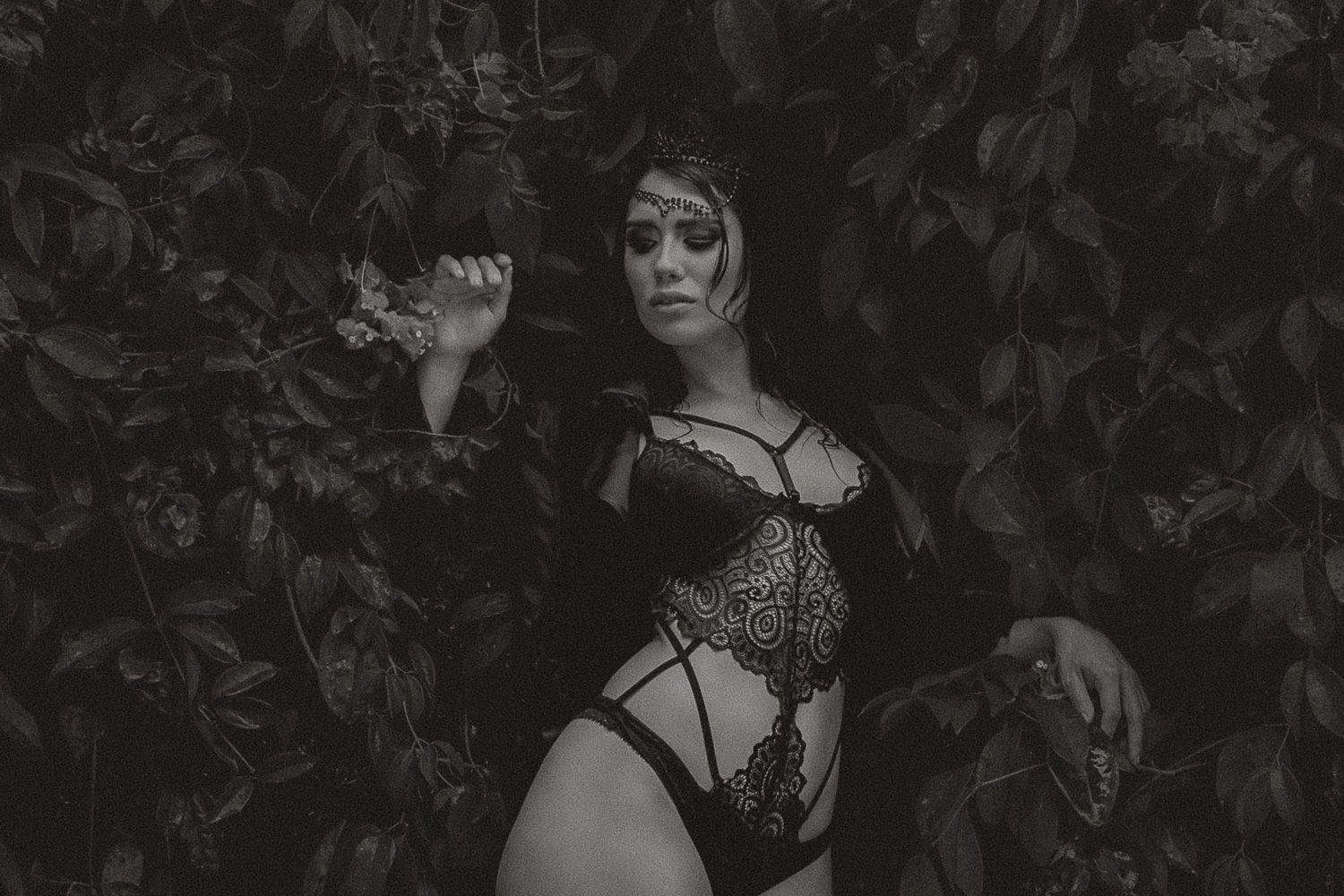 Black and white photo of a woman in lace lingerie posing among lush dark foliage.