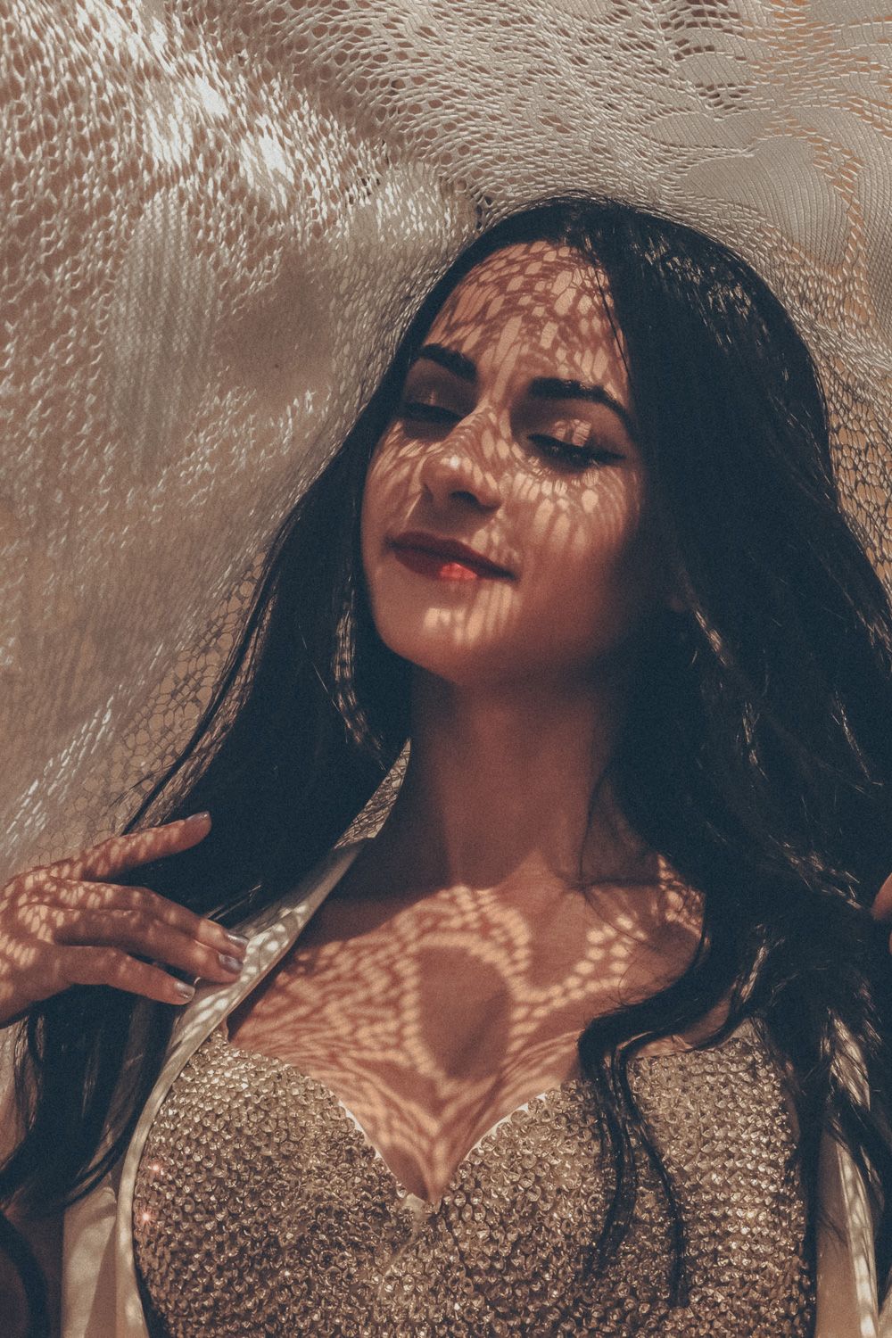 Woman with dark hair poses with eyes closed, dappled light patterns casting lace shadows on her face and décolletage.