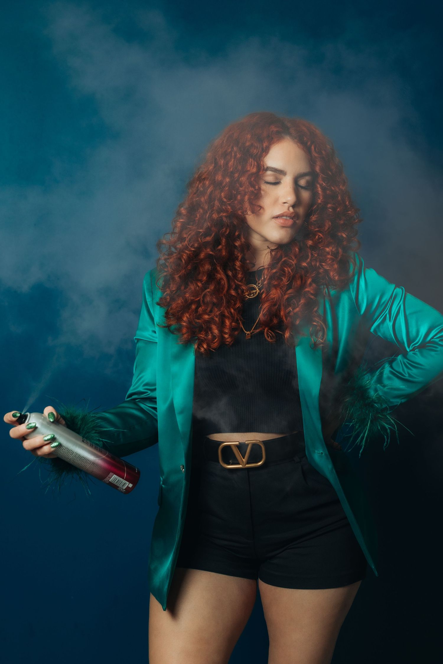Curly red-haired woman in teal blazer and black shorts holding a spray can with colorful smoke on dark background.