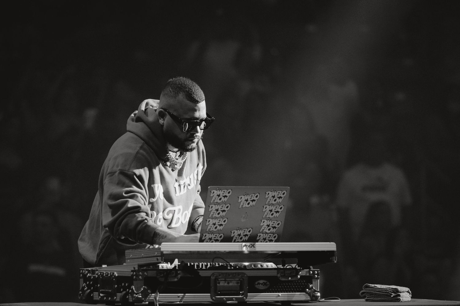 Black and white photo of a DJ performing on stage with equipment under dramatic lighting.