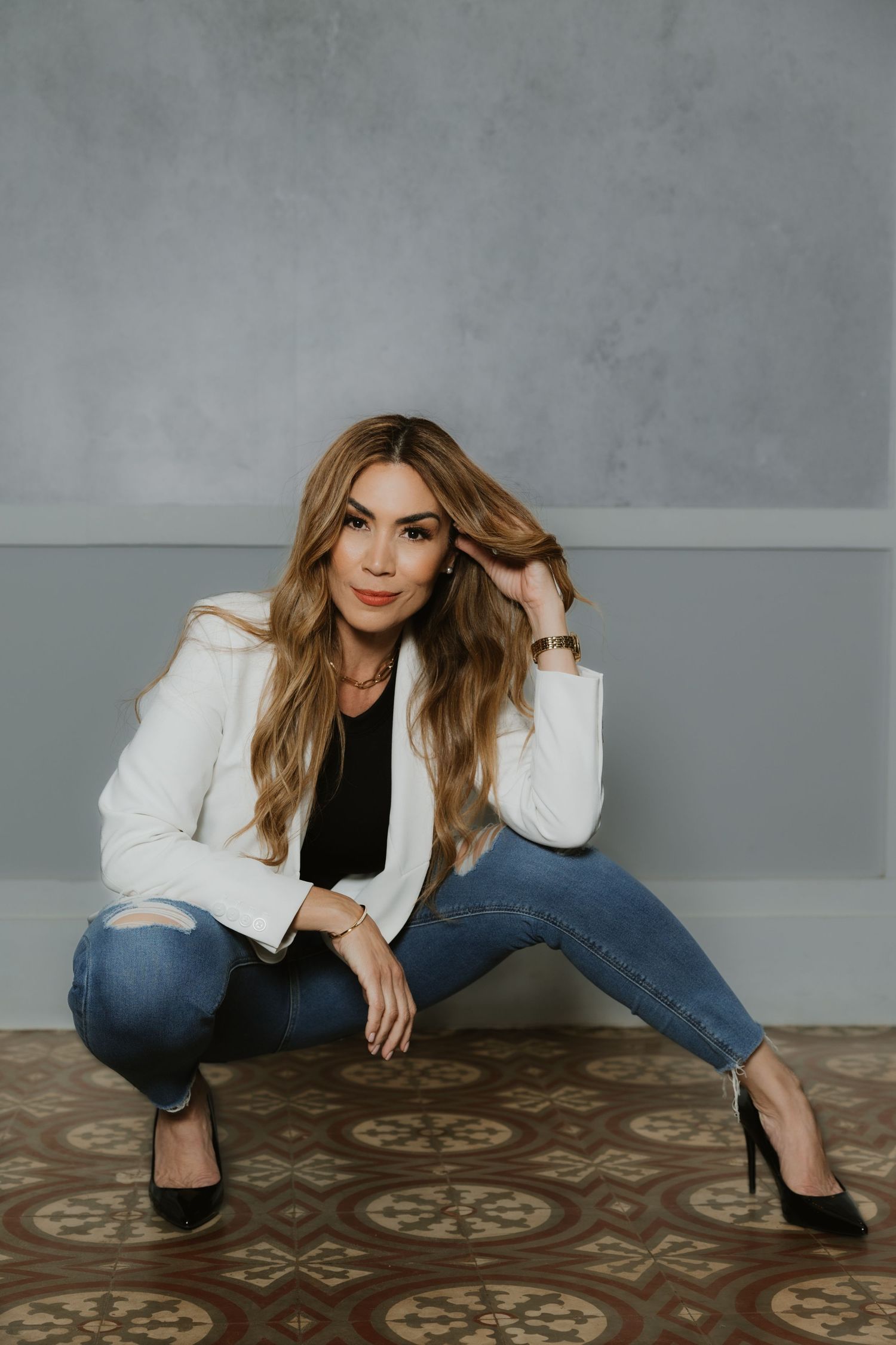 Woman in white blazer, black top, ripped jeans, and heels sitting on patterned floor against gray wall.