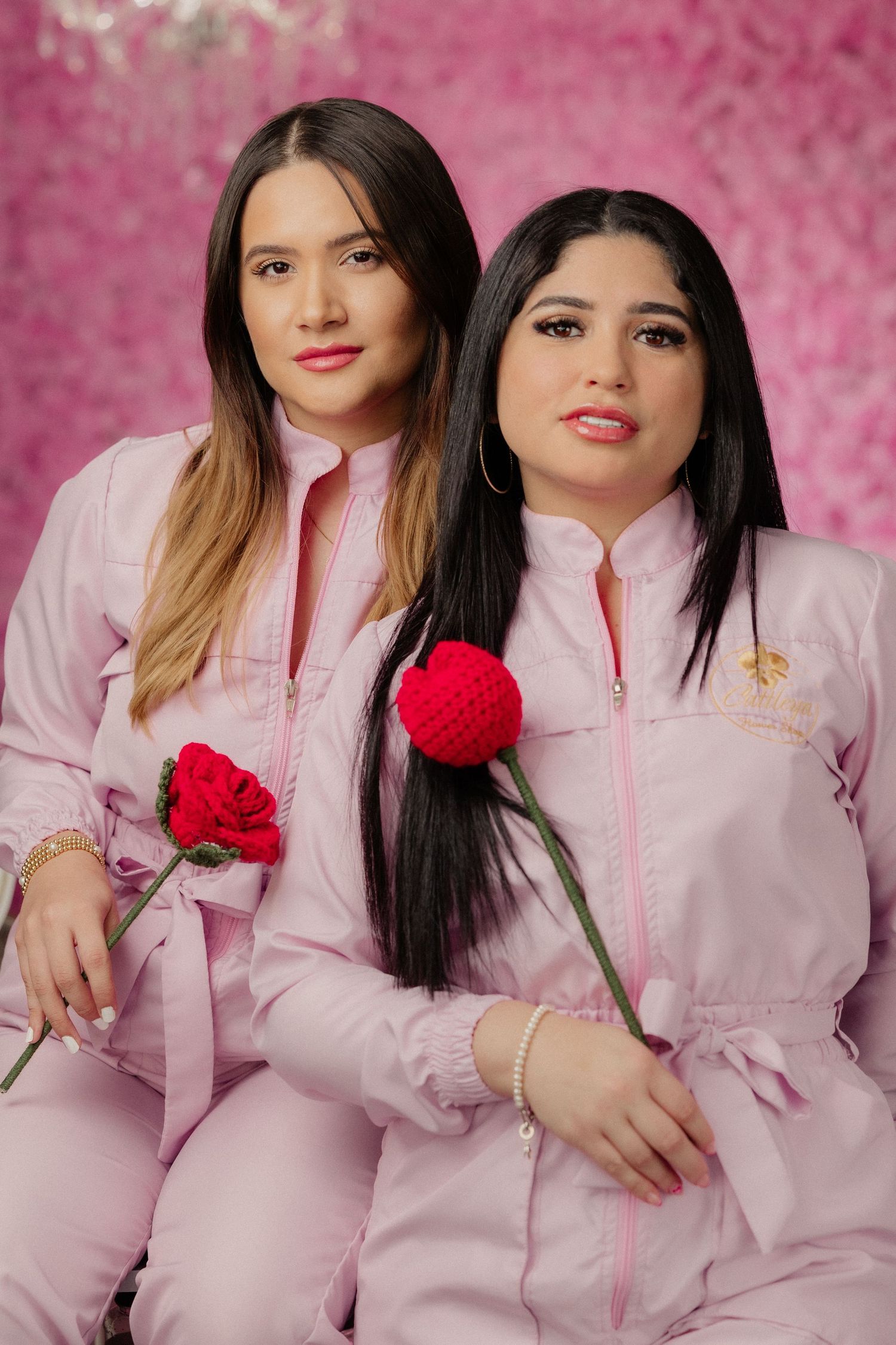 Two women in matching pink jumpsuits holding red roses pose together against a pink floral background.