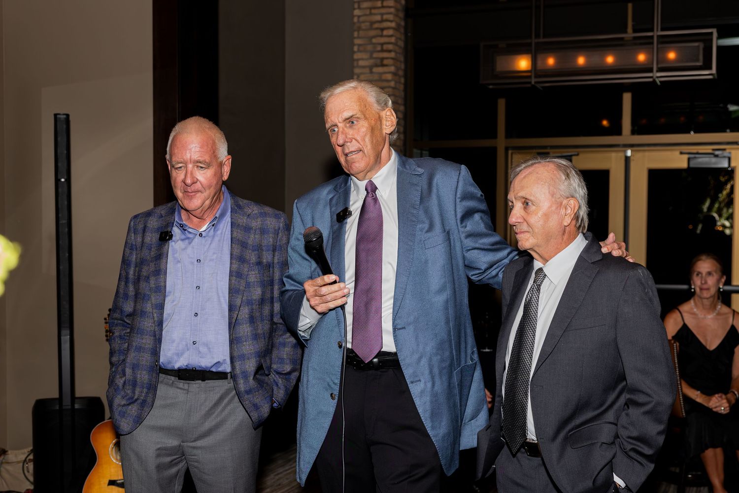 Three elderly men in formal attire stand together at an indoor event, with the center man holding a microphone.