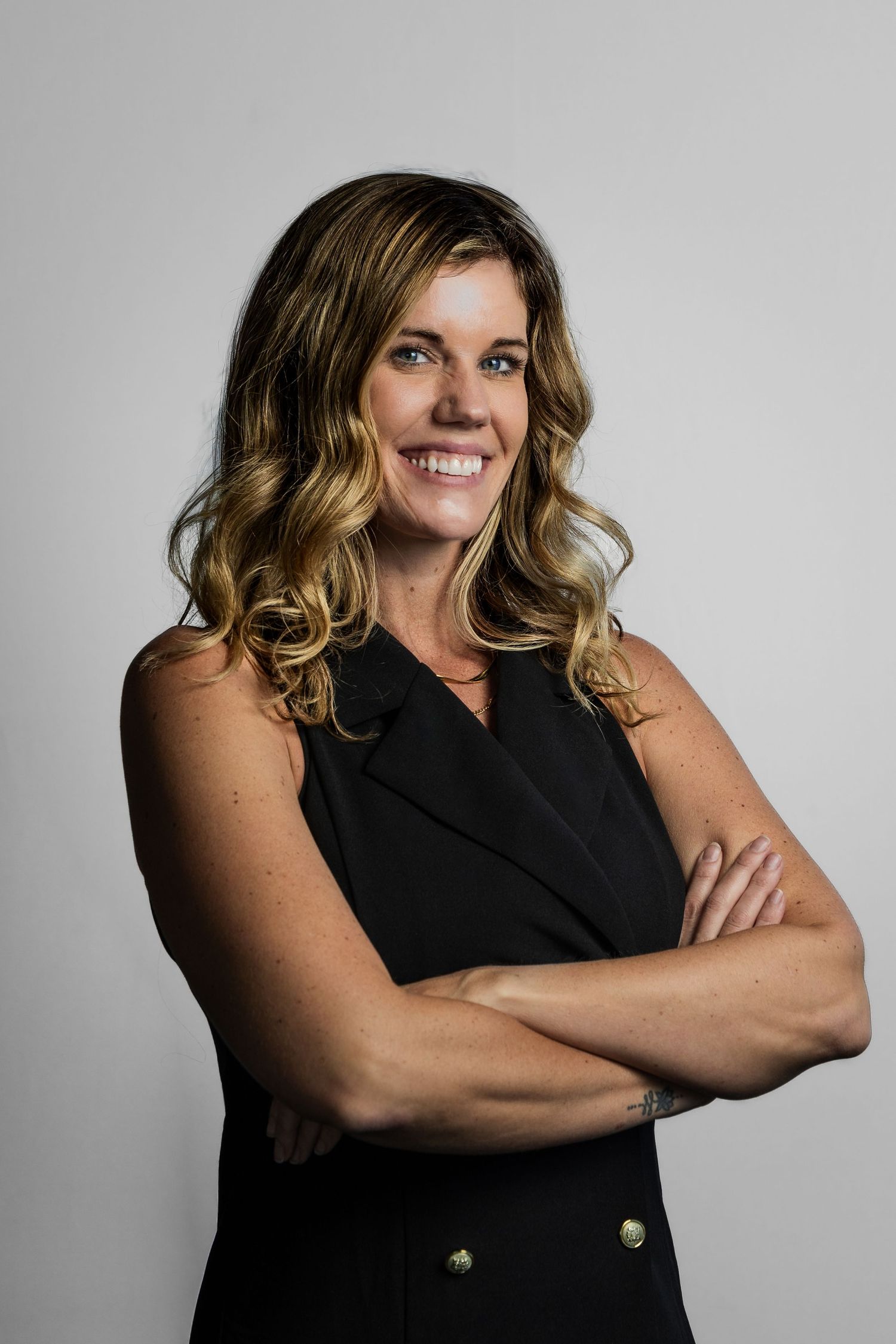 Smiling woman with wavy blonde hair in black sleeveless blazer, arms crossed, posing confidently against gray background.