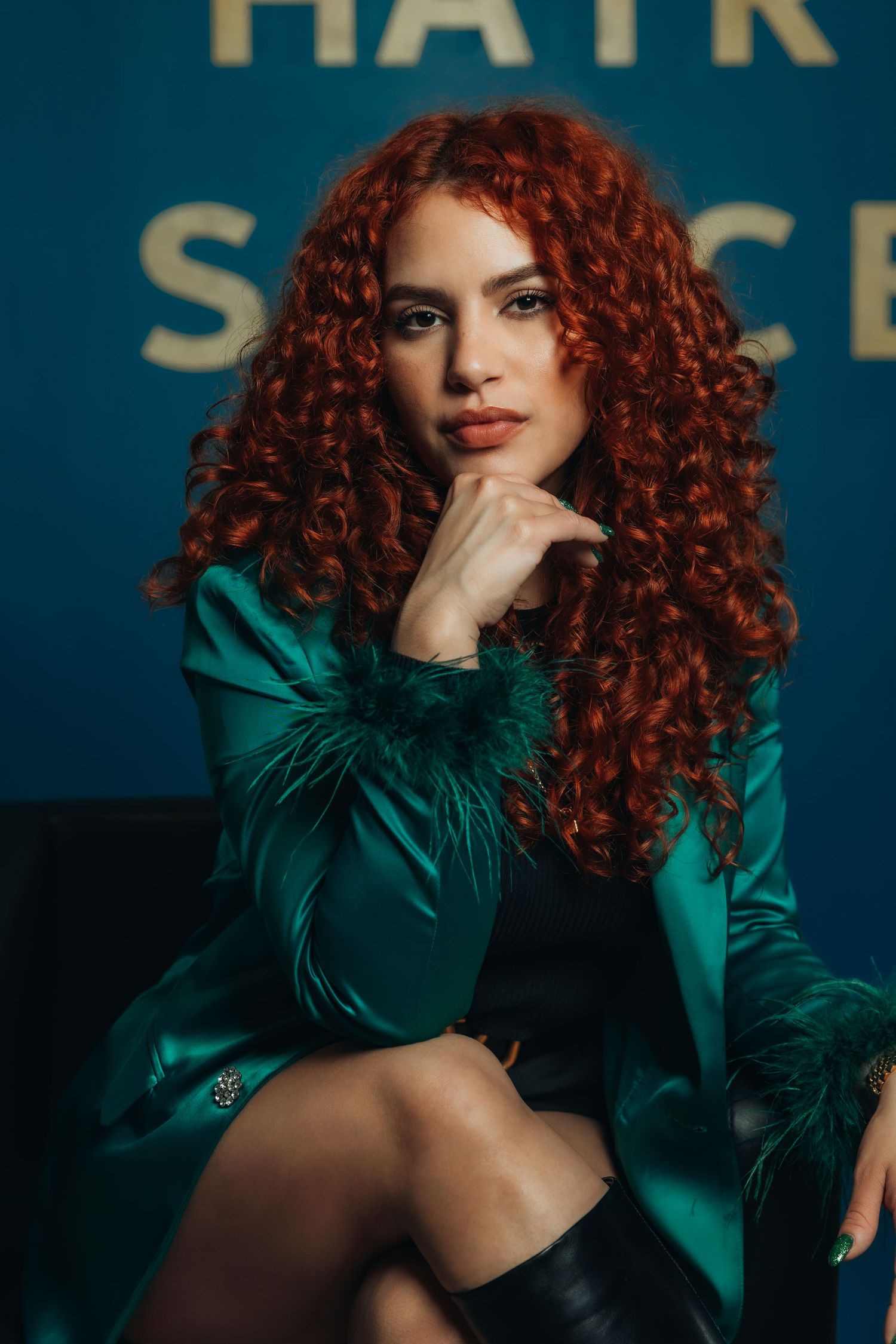 A woman with voluminous red curly hair poses in a teal satin jacket with feather trim against a blue backdrop.