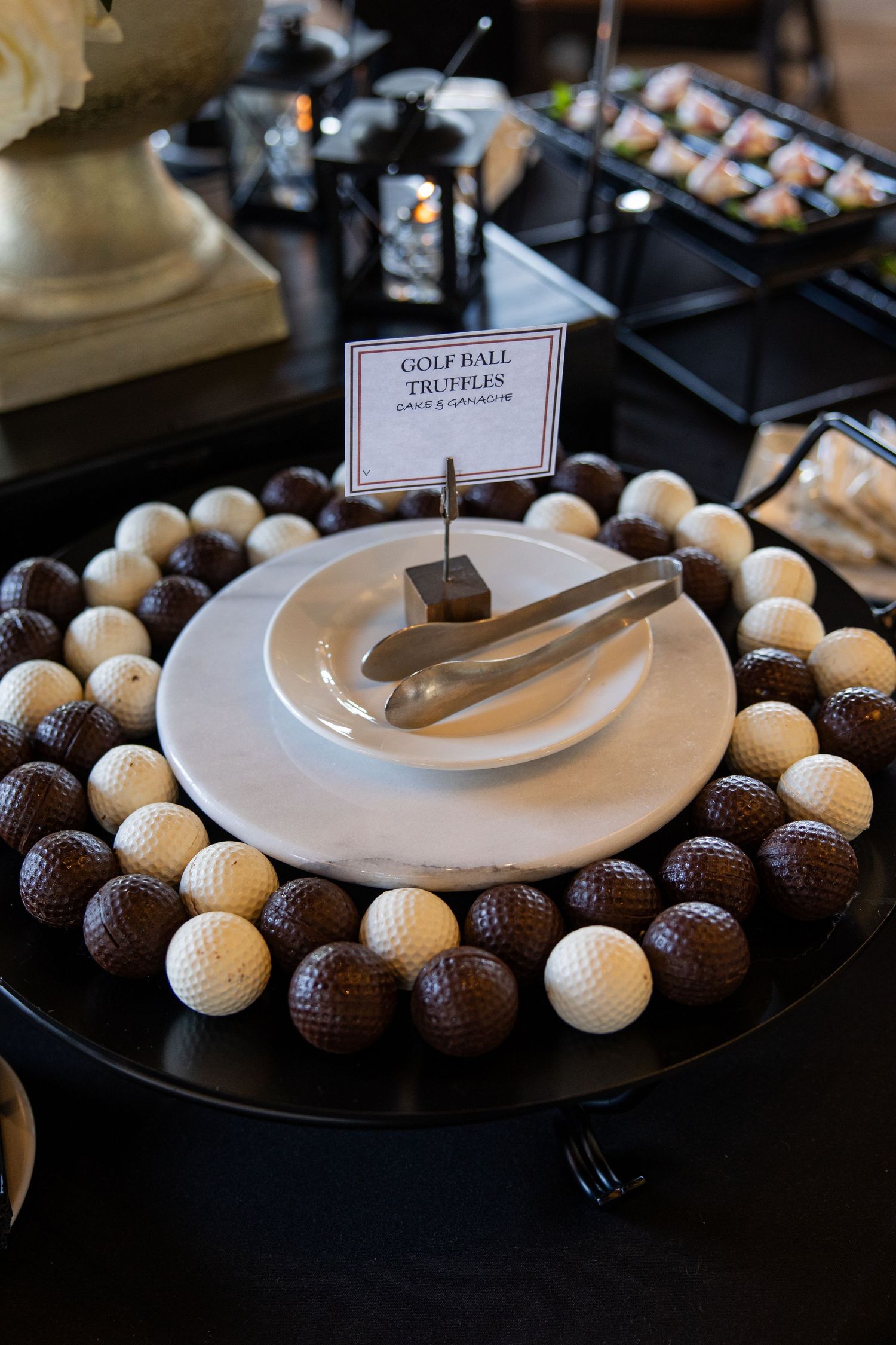 Golf ball truffles display with dark and white chocolate treats arranged around a white plate with tongs at a dessert table.