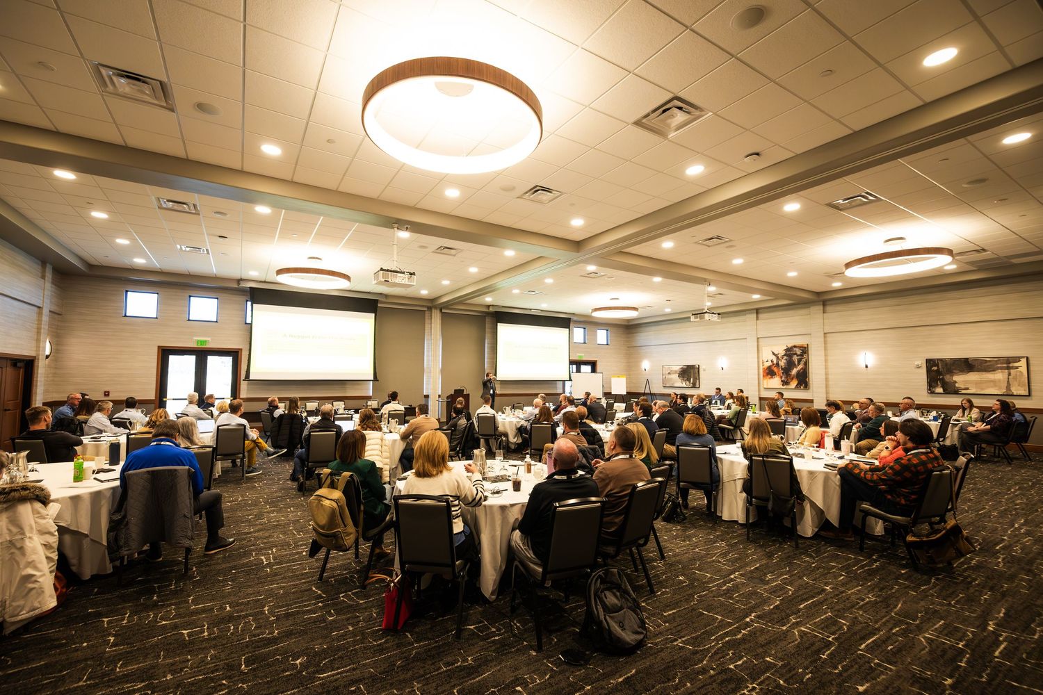 Large conference banquet hall filled with attendees seated at round tables during a professional event or seminar.