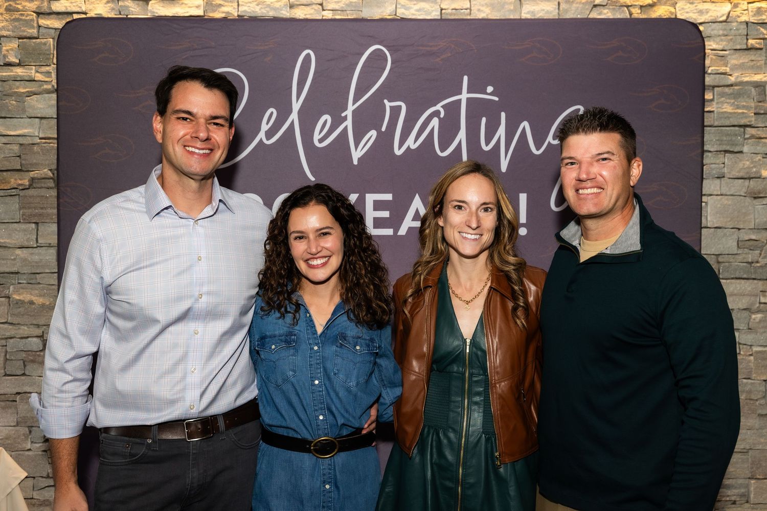 Four smiling adults pose together in front of a celebratory sign at an indoor event.