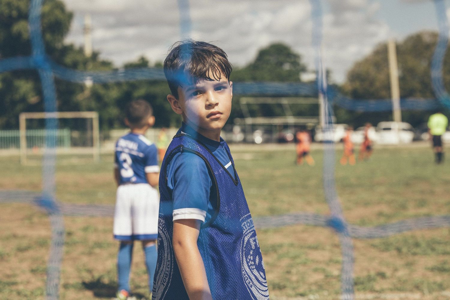 Young boy in blue soccer jersey standing on a football field during a youth soccer match outdoors.