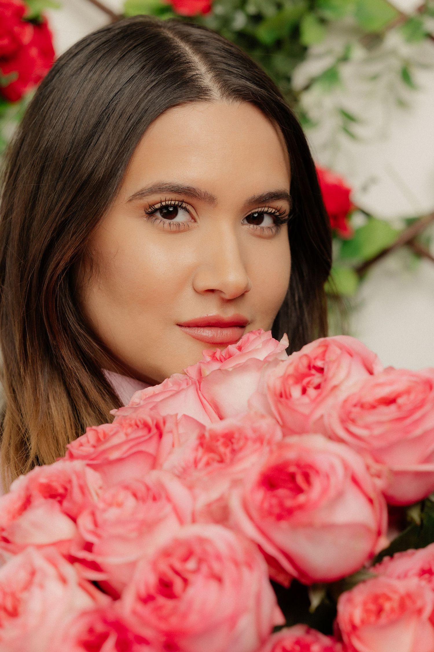 A young woman with dark hair holds a large bouquet of pink roses near her face, with red flowers in the background.