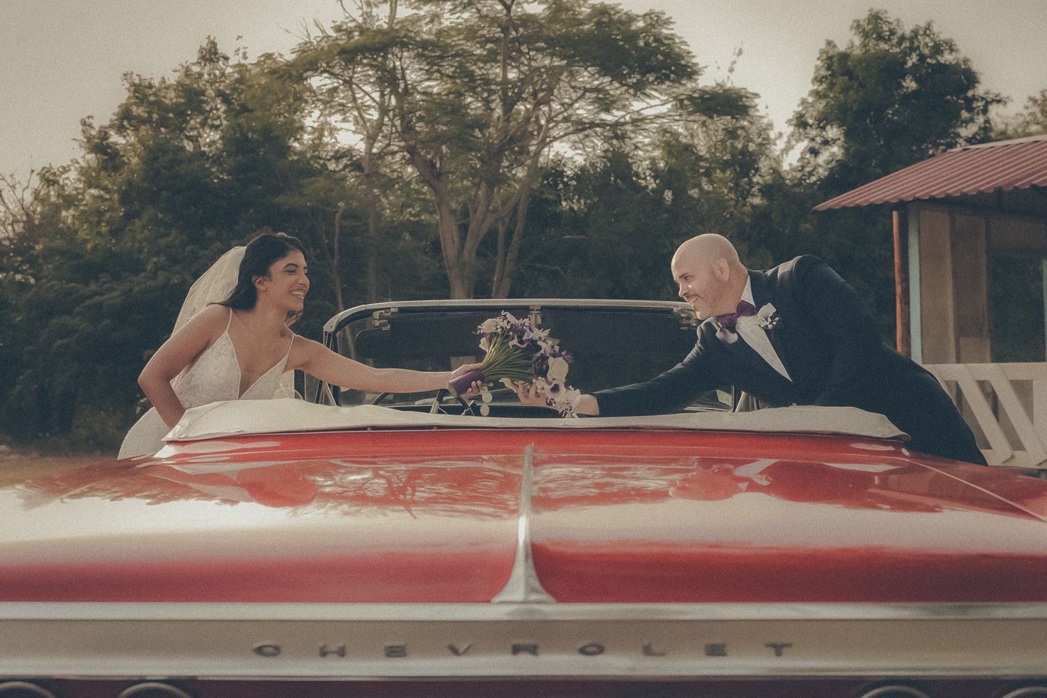 Bride and groom exchanging flowers across a red vintage Chevrolet convertible