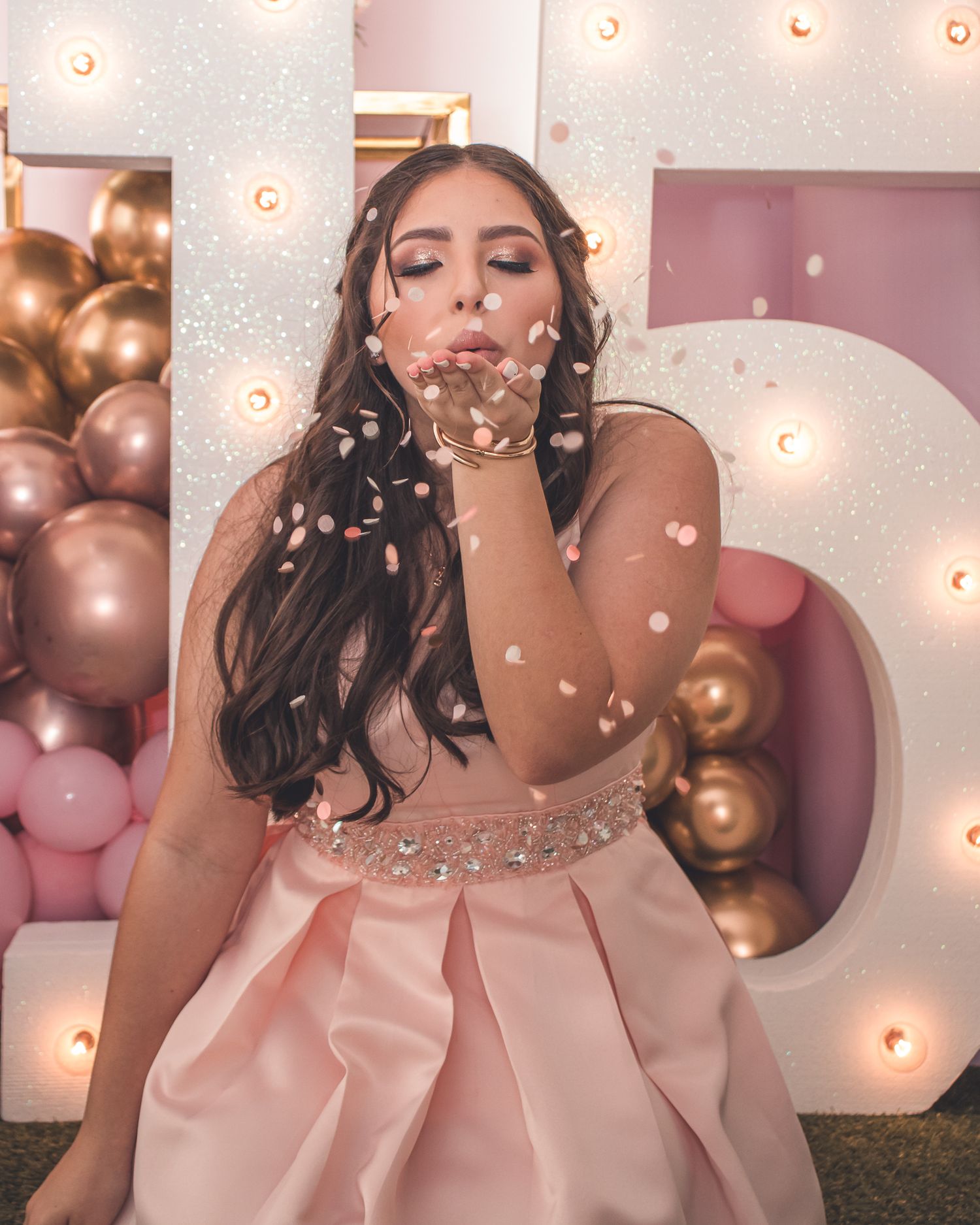 Birthday portrait of a teenage woman blowing confetti toward the camera surrounded by pink and gold balloons